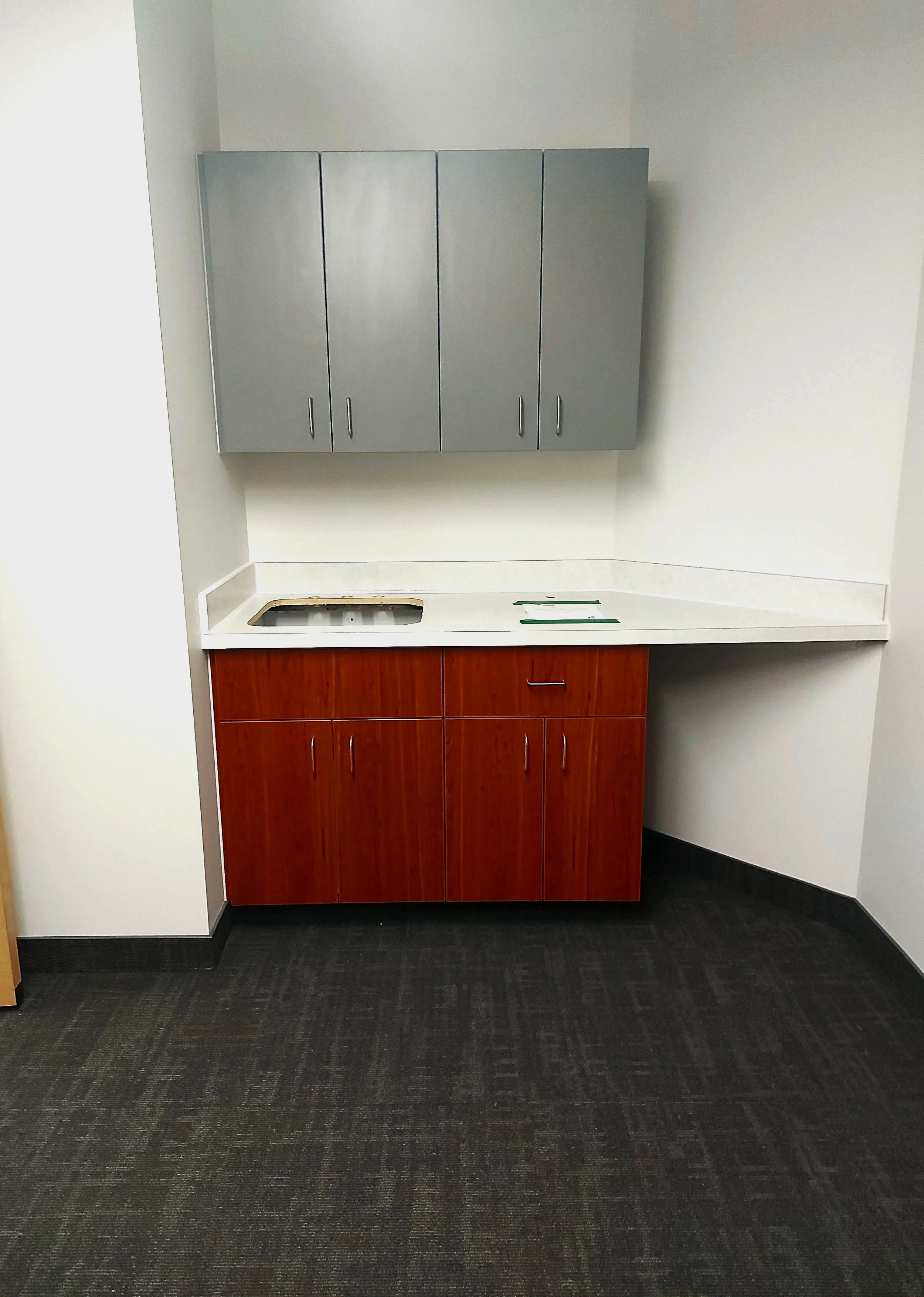 Empty office break area with upper gray cabinets, lower wooden cabinets, a countertop, and carpeted flooring.