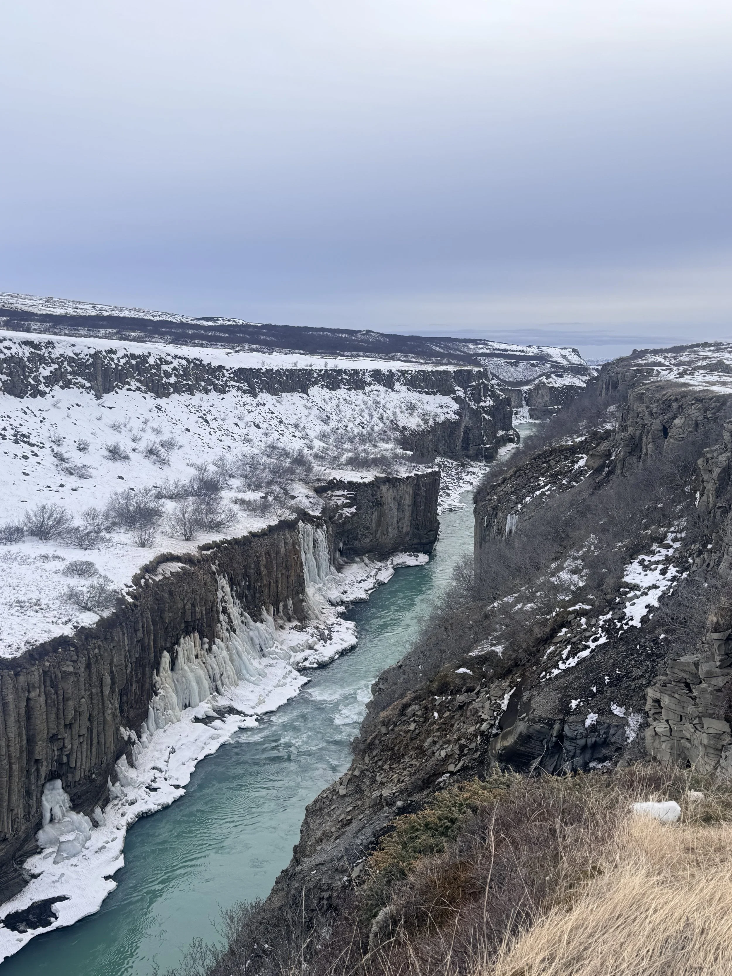 A river flowing through a snow-covered canyon with cliffs on either side, under a cloudy sky.