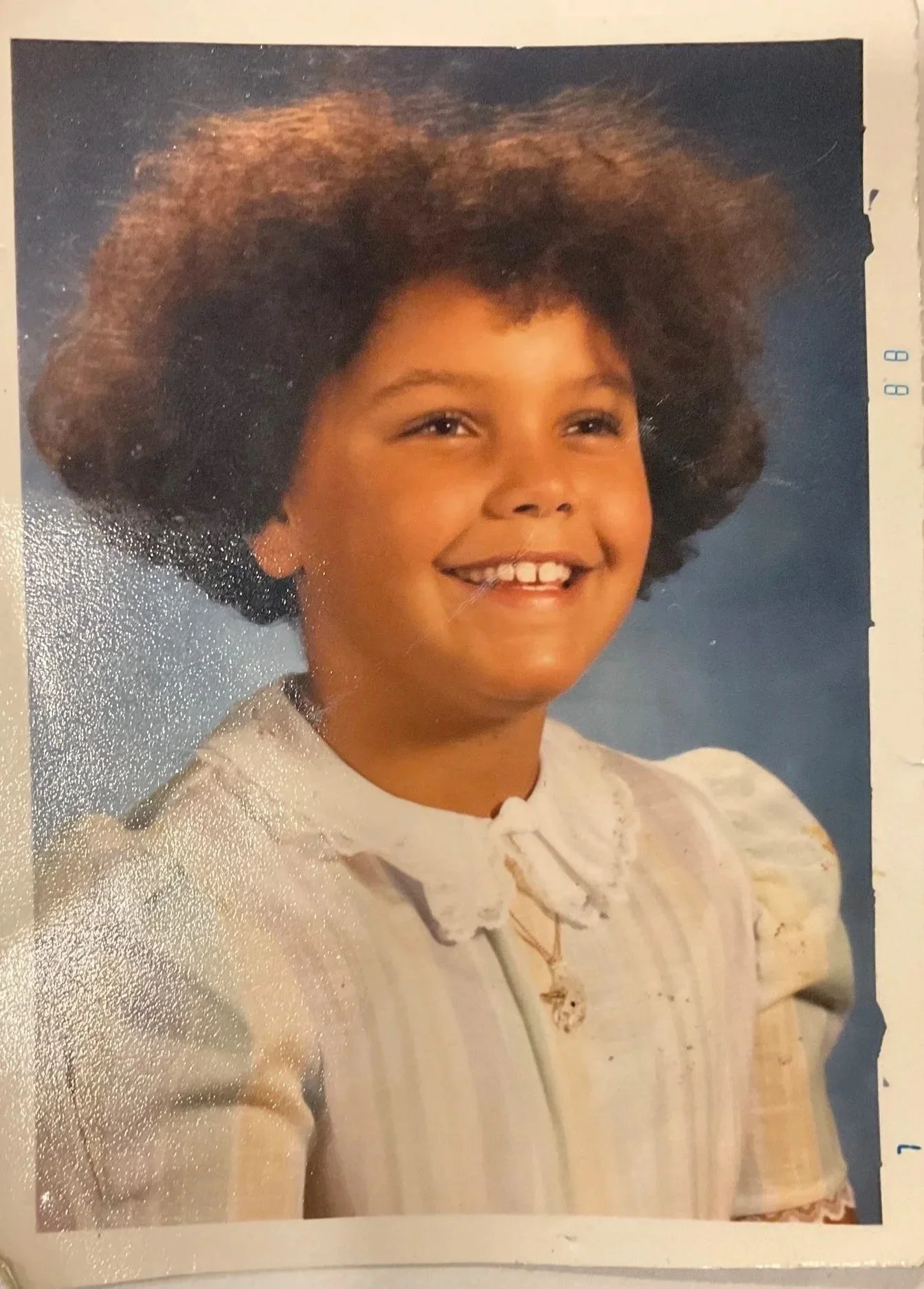 A young girl with a big, curly afro hairstyle, smiling, wearing a beige top with lace details, and a small pendant necklace, against a blue background.