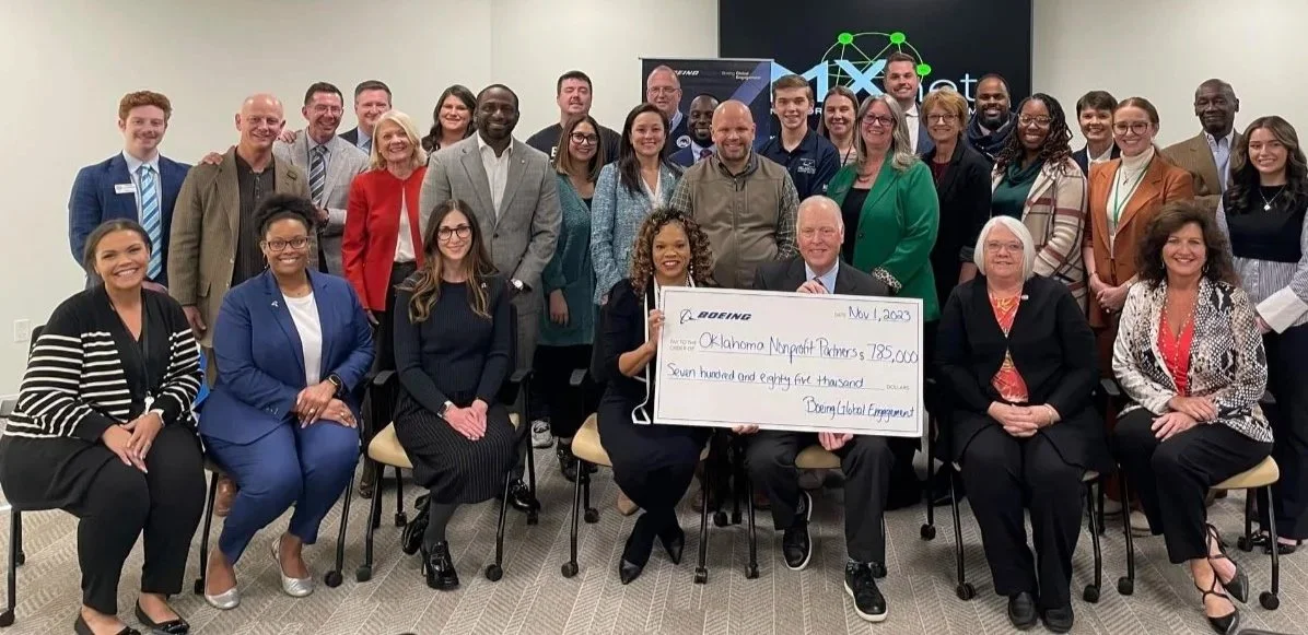Group of diverse professionals gathered in a conference room for a photo. Several individuals are seated in the front row, holding a large check made out to Oklahoma Momentum Partners for $725,000, dated November 1, 2020, related to alcohol engagement. Others stand behind them, smiling for the picture, with a presentation screen or banner in the background.