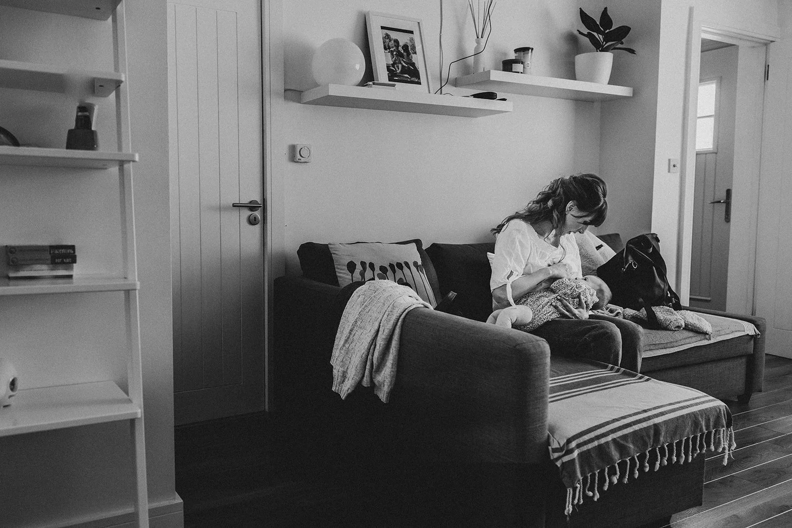 A black-and-white photo of a woman sitting on a sofa, holding a baby, in a cozy living room with shelves, framed pictures, and houseplants.