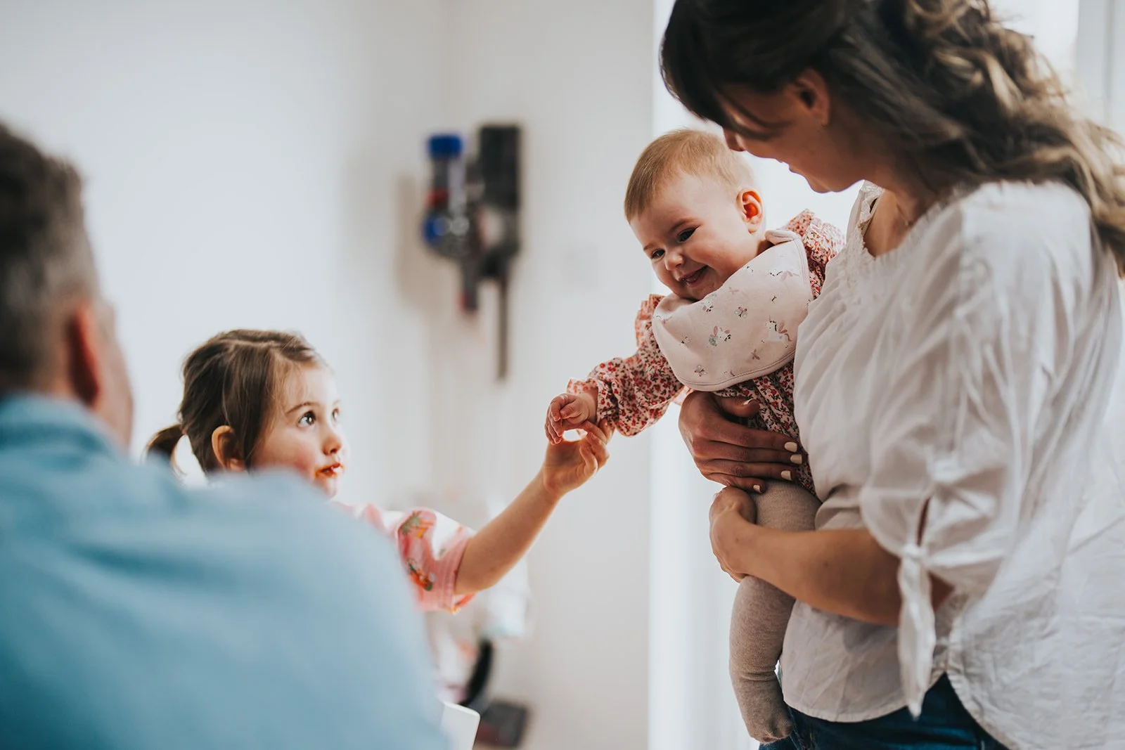 A woman holding a smiling baby girl in a hospital room, reaching out to a young girl and an out-of-focus man, possibly her father, who are looking at the baby and reaching to hold her hand.