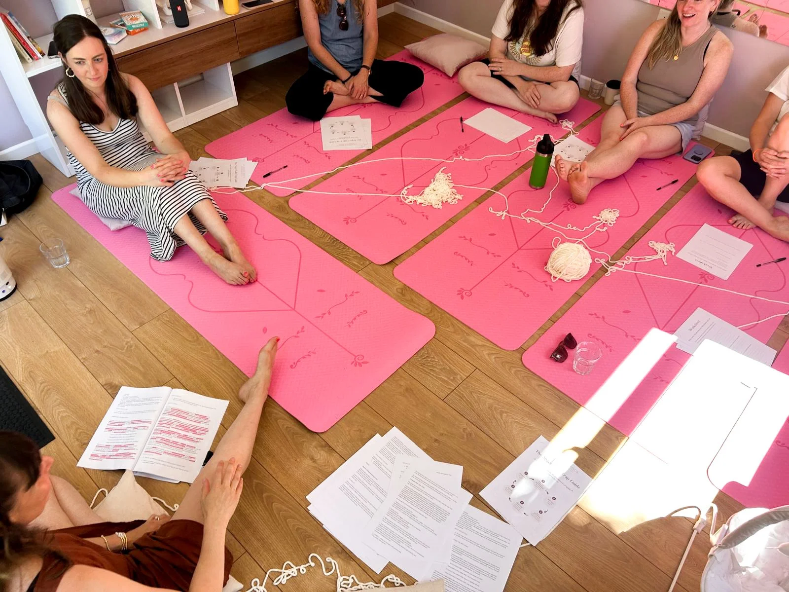 A group of women sitting on pink yoga mats in a circle, participating in a workshop or discussion, with papers, pens, and yarn scattered around.