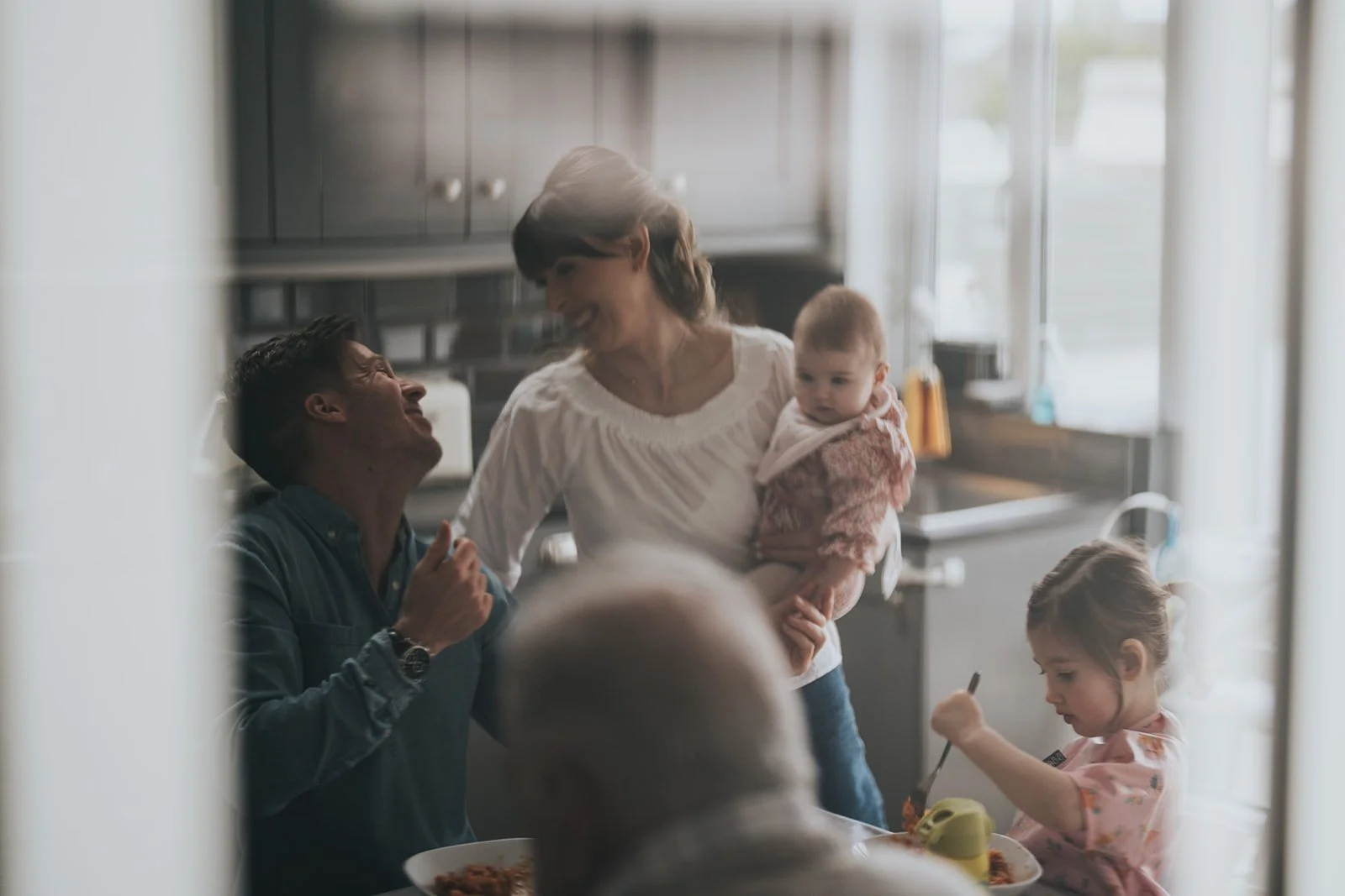 A family in a kitchen, smiling and interacting. A woman holds a young child while talking to a man, a girl eats at the table, and other family members are present.