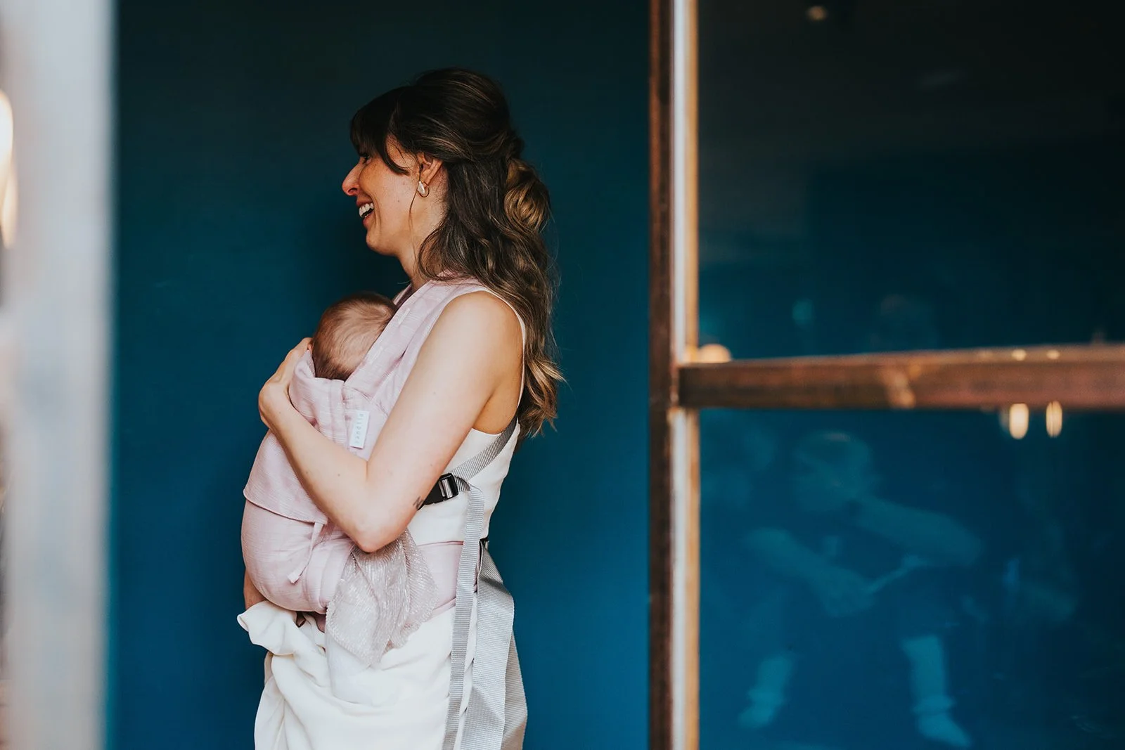 A woman with long brown hair smiling while holding a baby wrapped in pink cloth against a dark blue wall.