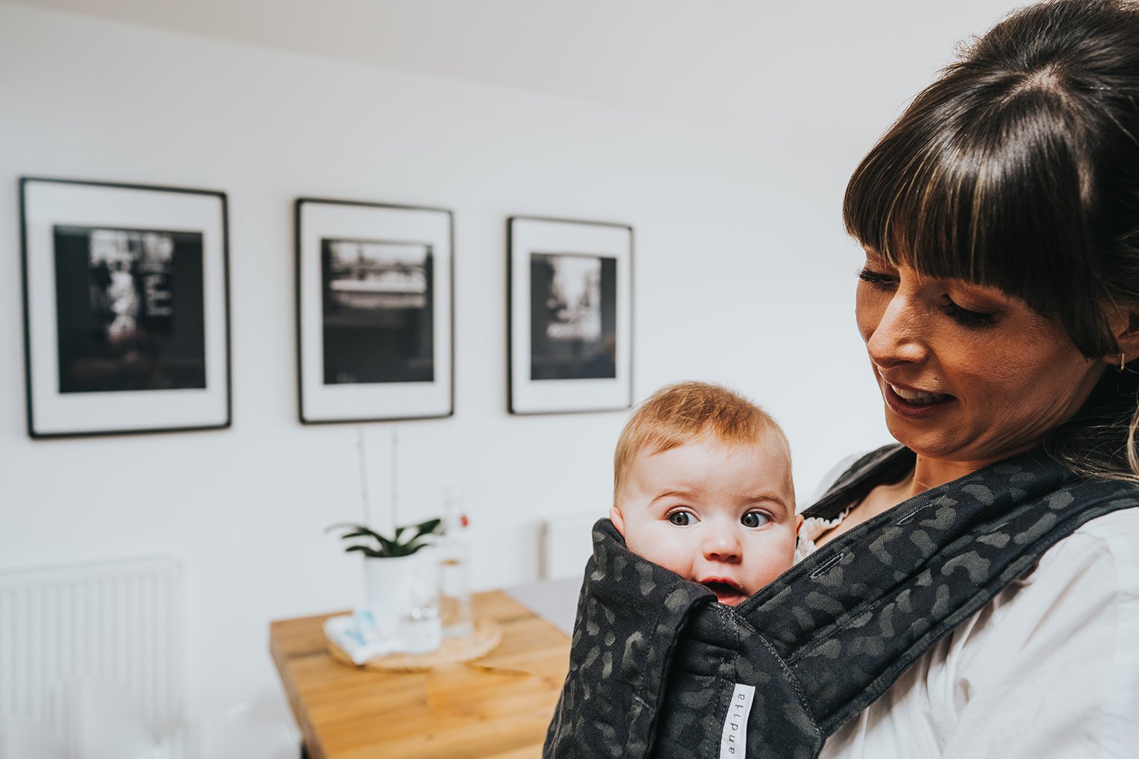 A woman with short dark hair smiling while carrying a baby in a black baby carrier. The baby has light hair and wide eyes. In the background, there are three framed black-and-white photographs on a white wall and a wooden table with a potted plant.
