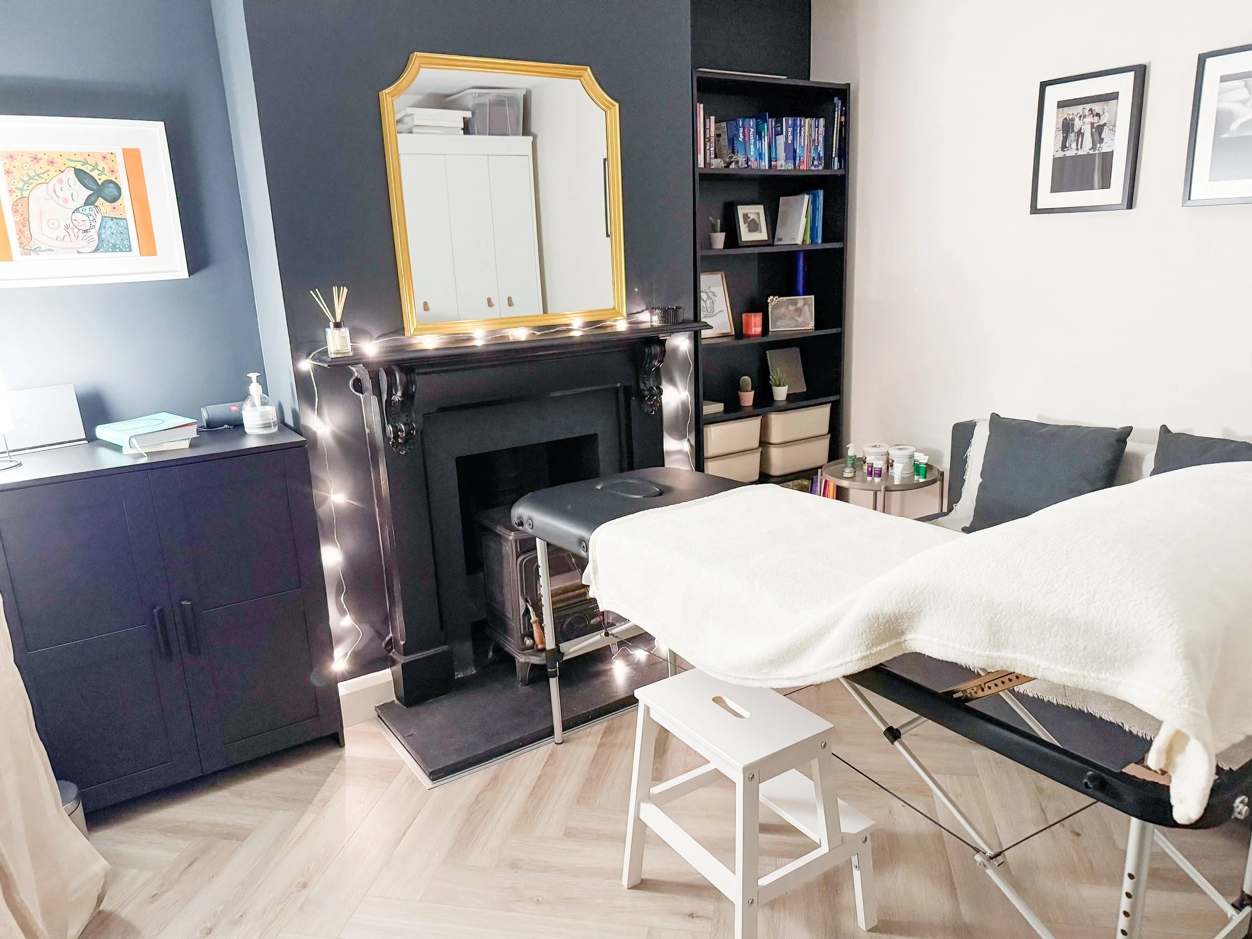 A clinical room with a massage or treatment table, a black fireplace mantel decorated with fairy lights, a large mirror with a gold frame, a black bookshelf filled with books and framed photos, and framed artwork on the walls.