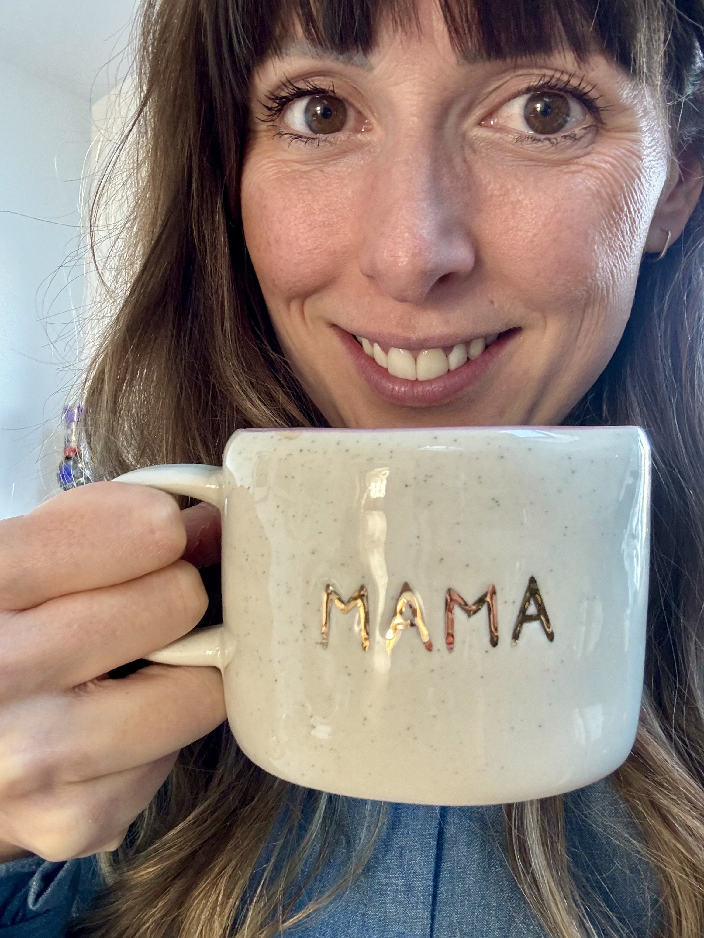A woman with long brown hair and a wide smile holding a ceramic mug that says 'MAMA' in multicolored letters, with a blurred background.