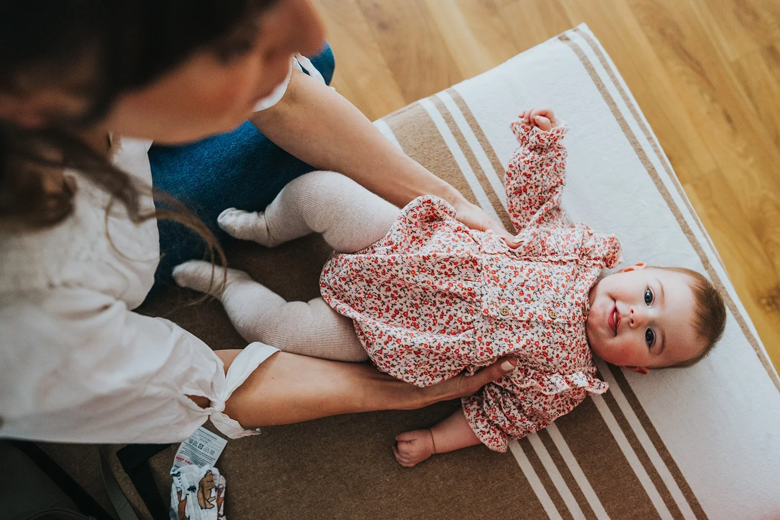 A young woman is gently holding a smiling toddler girl who is lying on a bench, wearing a floral dress and beige tights, with a wooden floor in the background.
