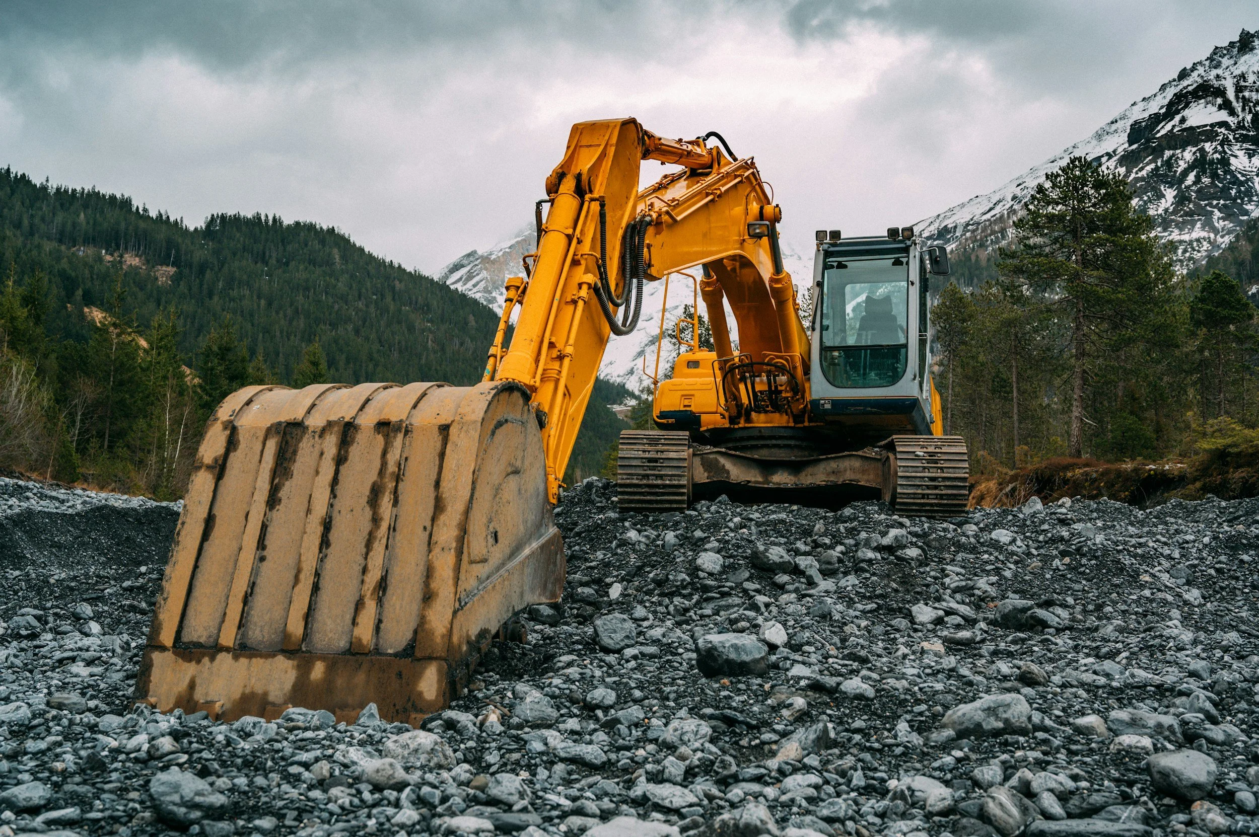 A yellow excavator working on rocky terrain with snow-capped mountains and pine trees in the background.