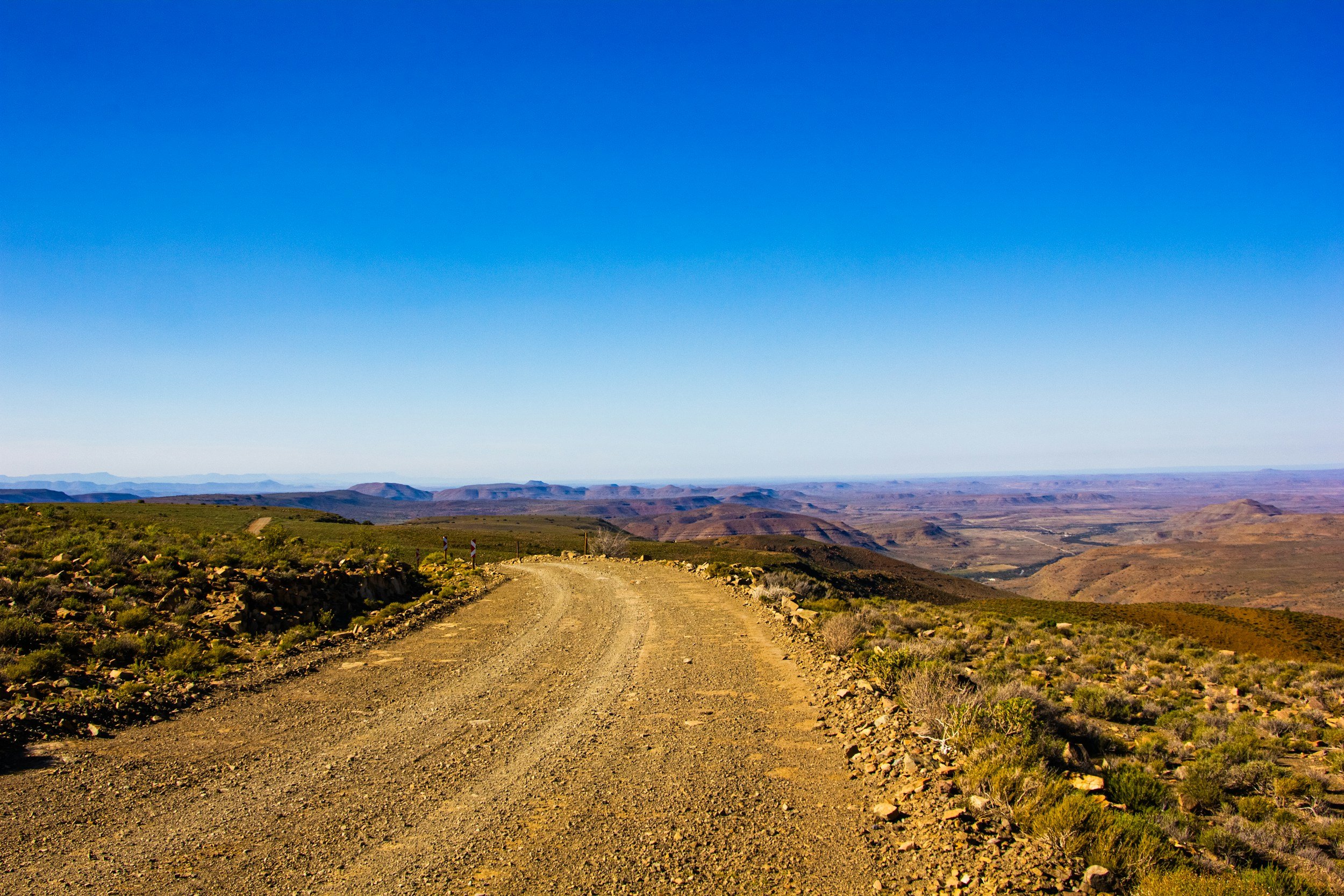 Dirt road in a desert landscape with green shrubs, distant mountains, and a clear blue sky.