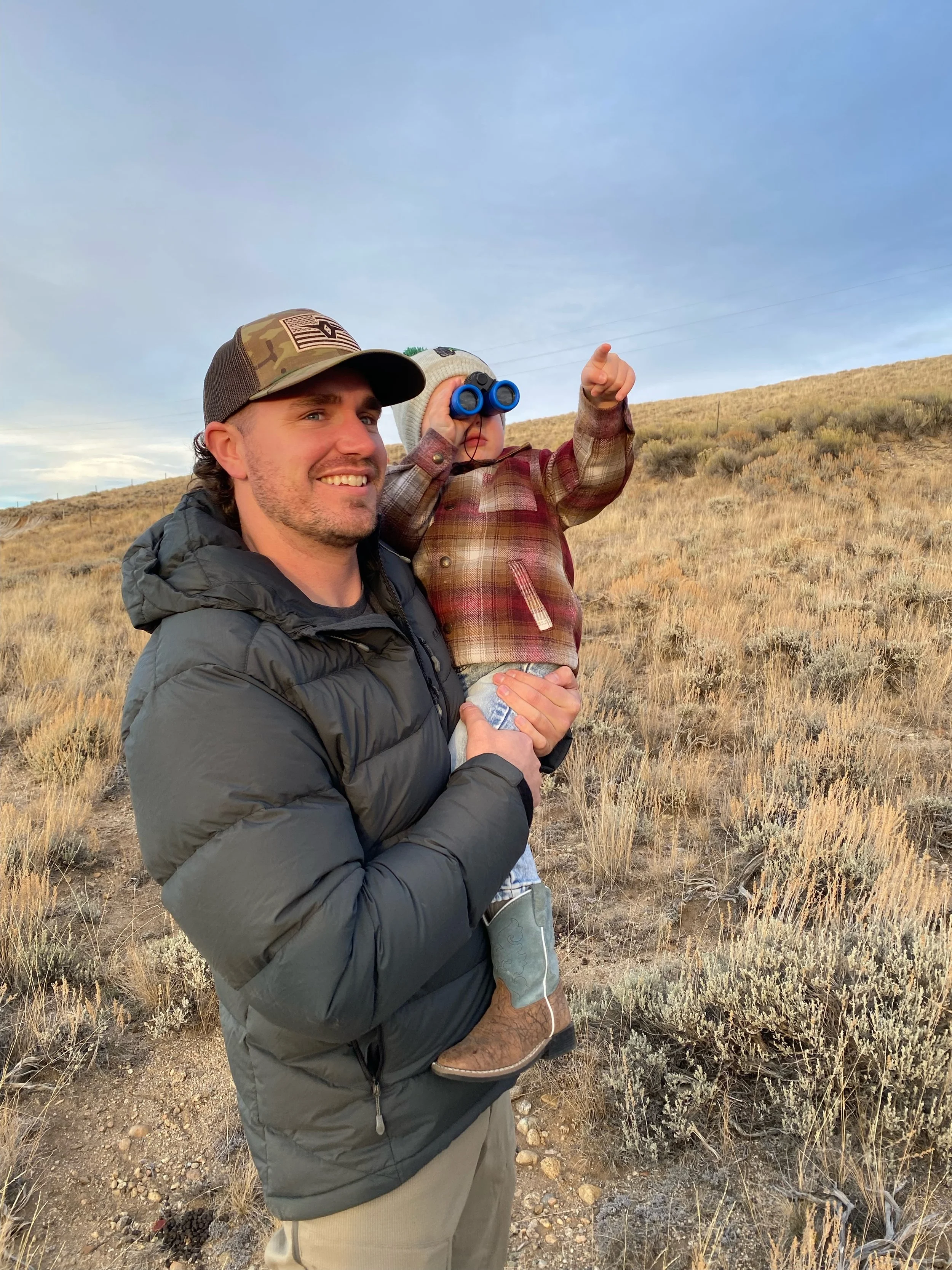 A man is holding a young child in a desert landscape with dry grass and shrubs. The man is wearing a black jacket and a camouflage cap. The child is wearing a plaid shirt, gray hat, and binoculars, pointing towards the sky.