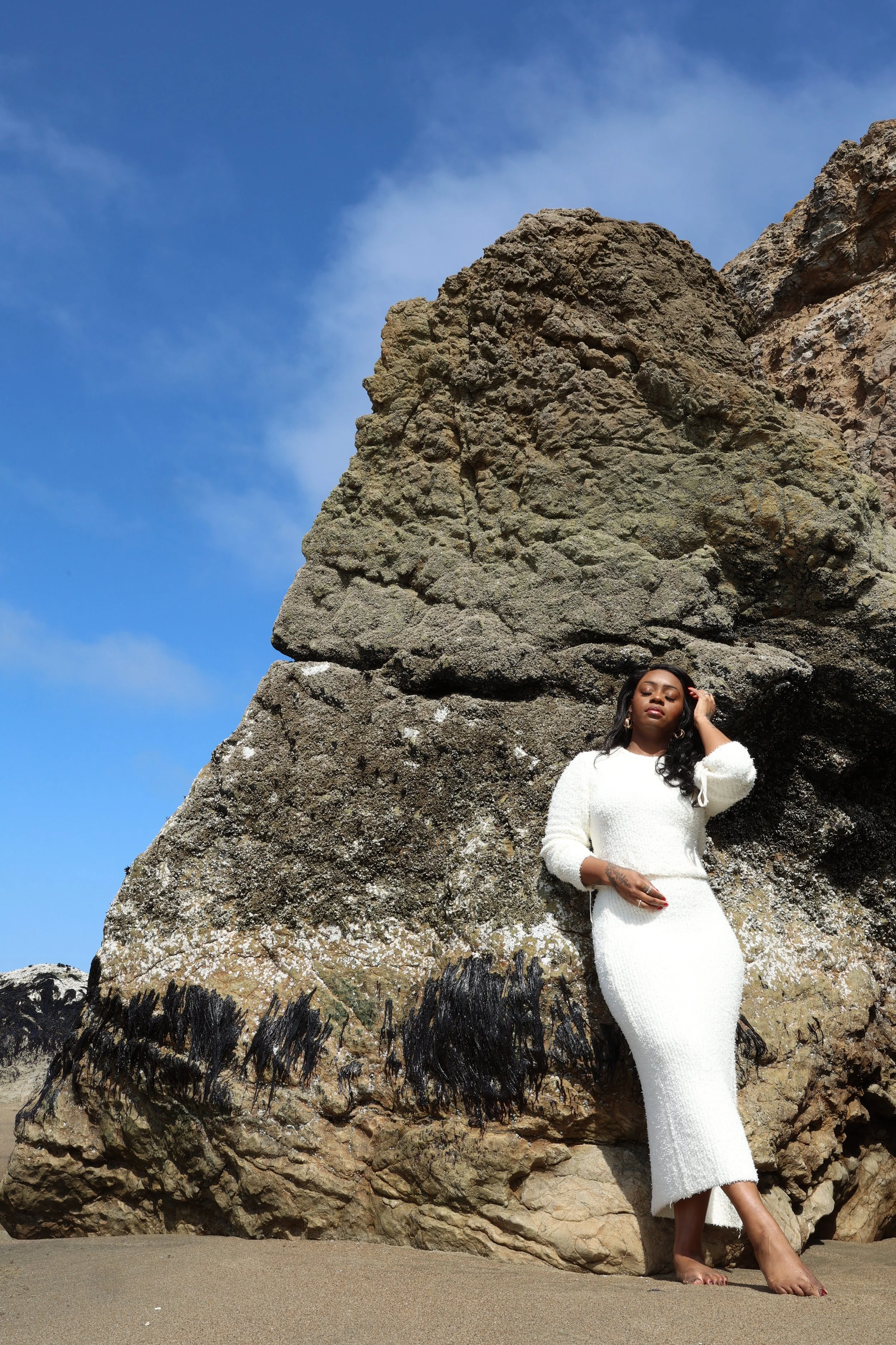 A woman in a white dress standing barefoot on a sandy beach, leaning against large volcanic rocks with a blue sky and some clouds in the background.