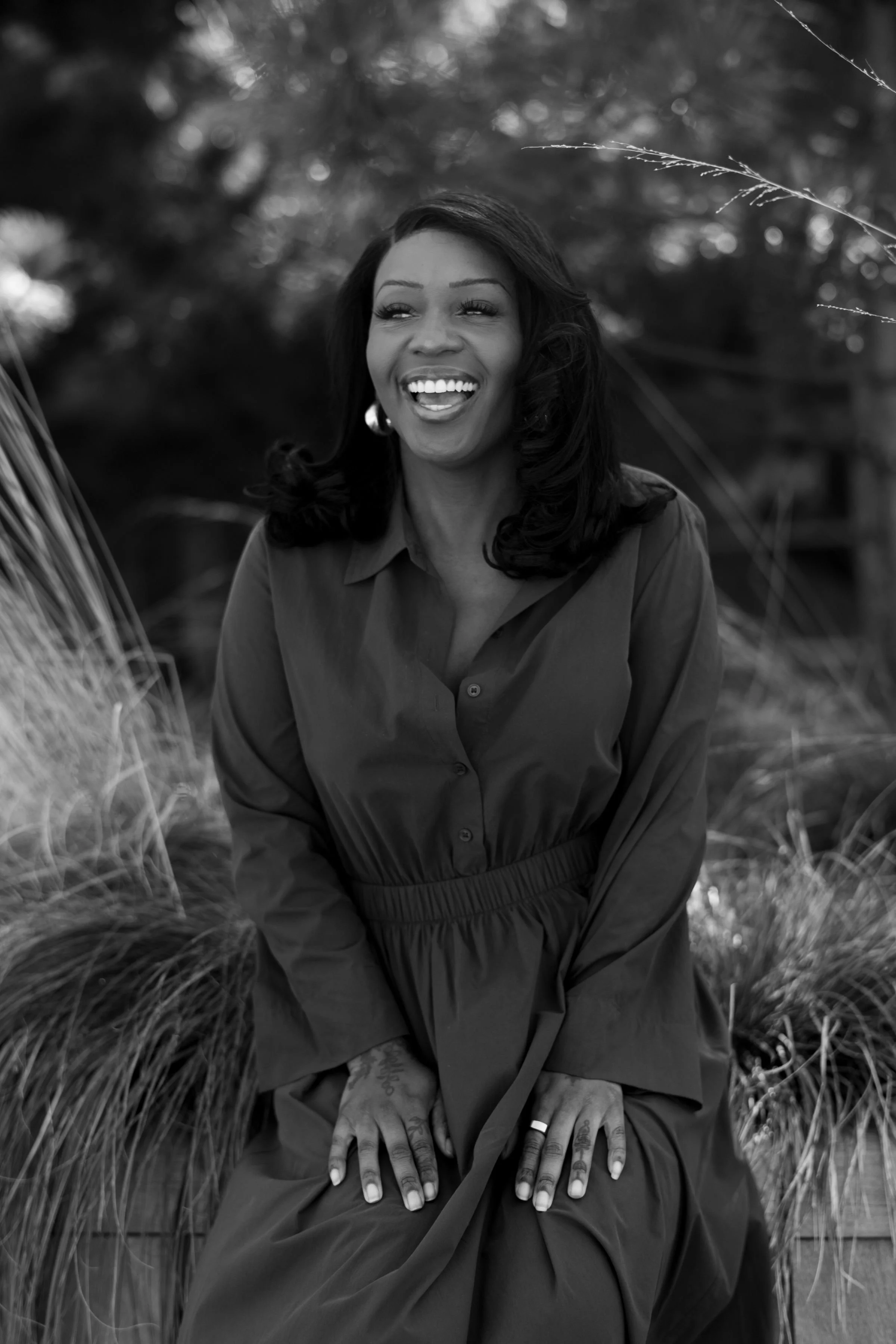 A woman with dark wavy hair smiling and looking to the side, sitting outdoors among tall grass and trees, wearing a dark, buttoned dress. Photo by Nadia Meli