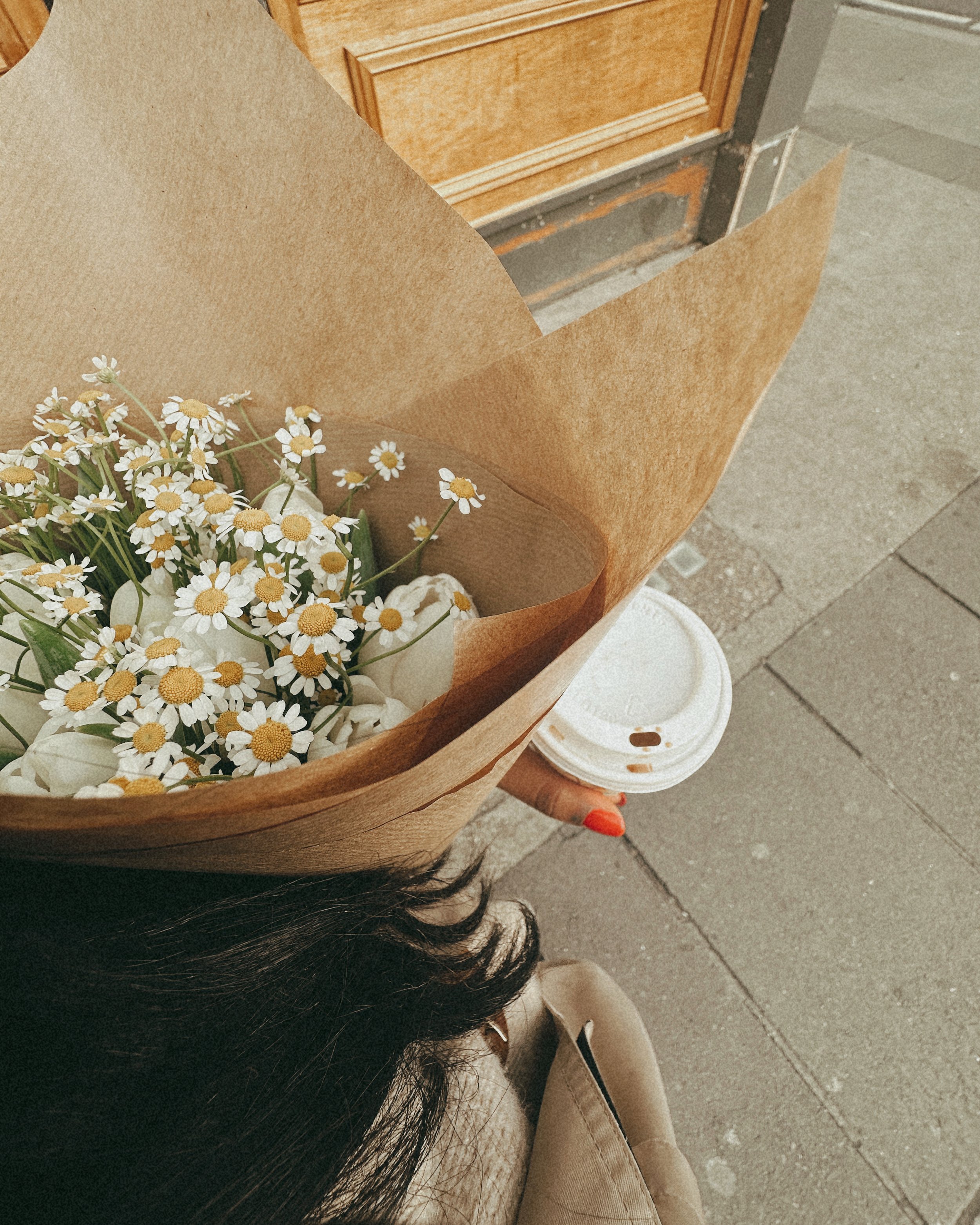 A person holding a paper coffee cup in their right hand, standing at a sidewalk near a wooden door, with a bouquet of daisies wrapped in brown paper in the foreground. The person is wearing beige pants and tan shoes, with dark hair visible at the bottom of the image.