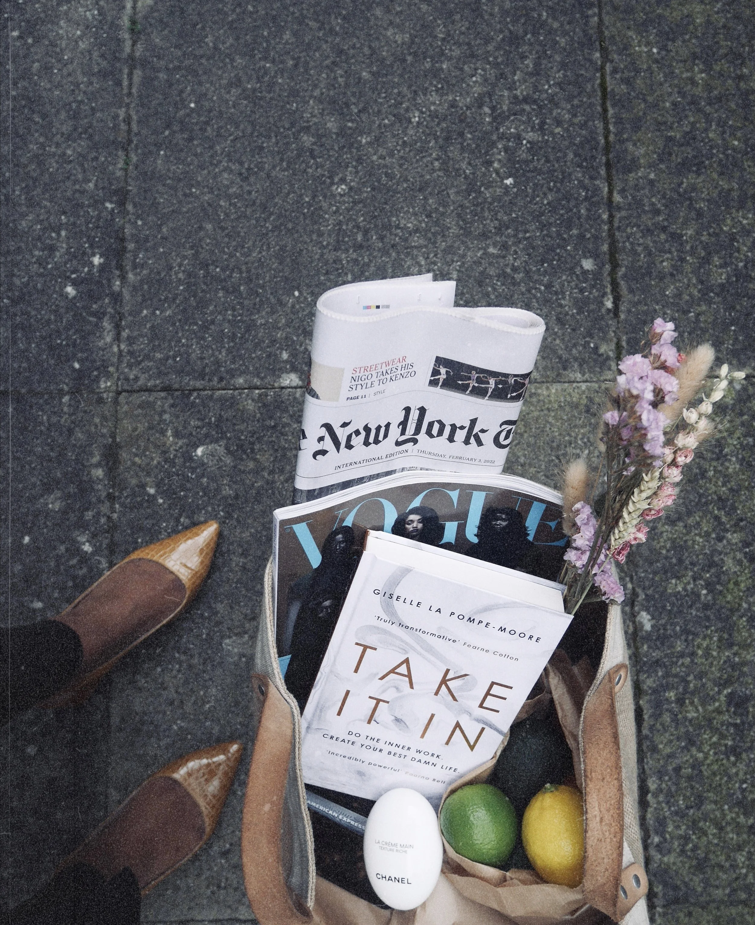 A person, likely a woman, wearing brown cowboy boots and jeans, stands on a concrete sidewalk next to a paper bag containing magazines, a white Chanel hand cream, a white egg, a green lime, and a yellow lemon, with a bouquet of pink flowers also in the bag.