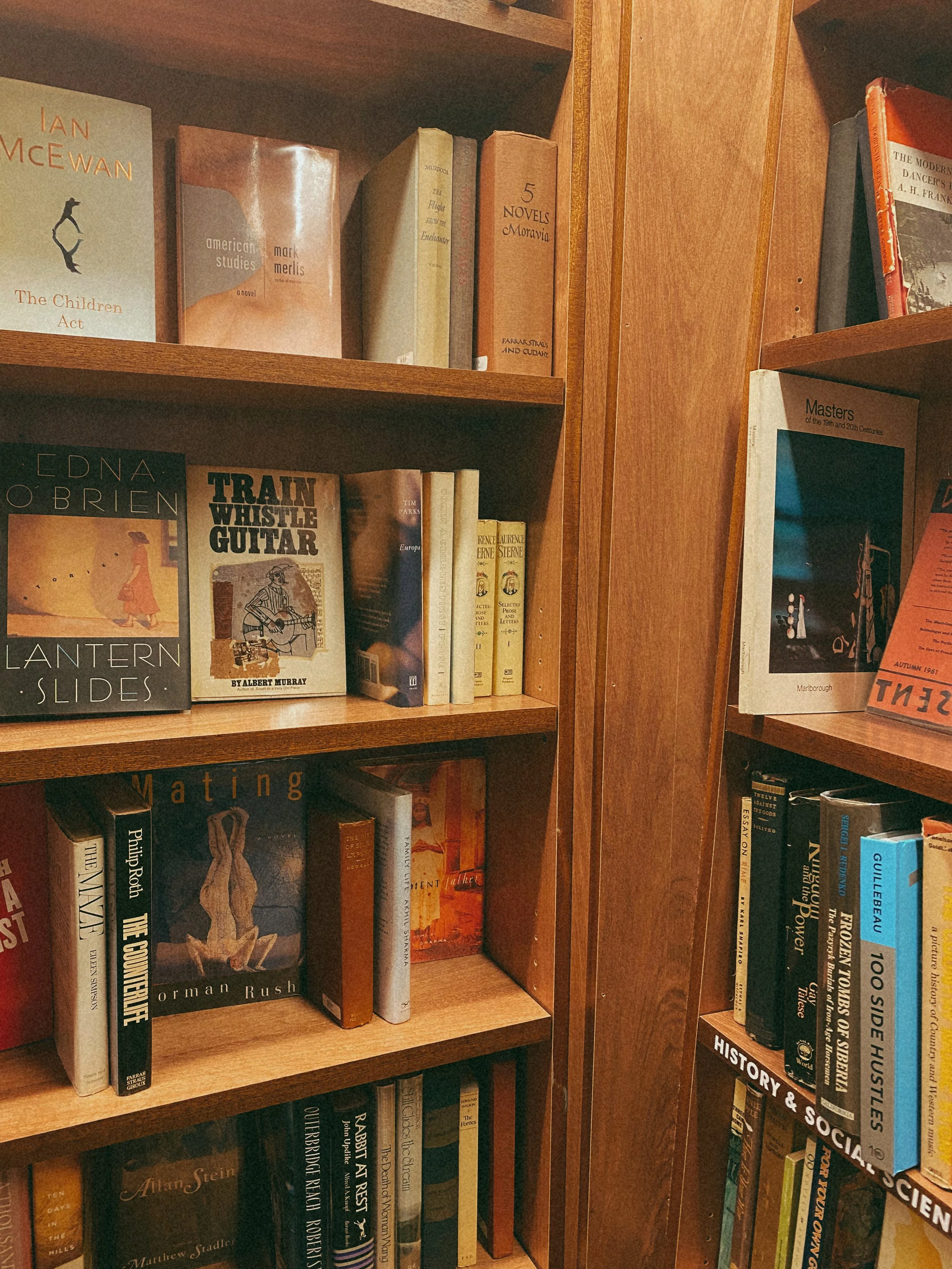 Bookshelf with multiple wooden shelves filled with various hardcover books, titles include 'The Children Act,' 'American Studies,' 'Train Whistle Guitar,' and 'Masters of the 19th and 20th Centuries.'