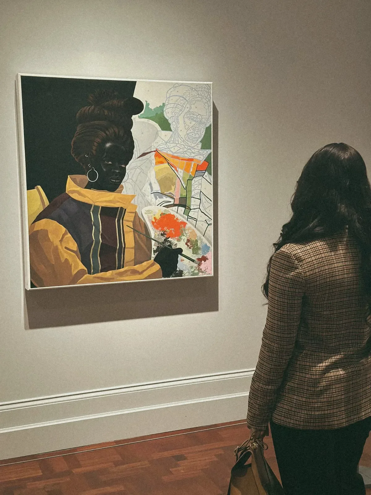 A woman with long dark hair wearing a brown plaid jacket is looking at a contemporary painting of a woman holding a palette and brush in an art gallery. The painting features a stylized portrait with vibrant colors and outlined sketches.