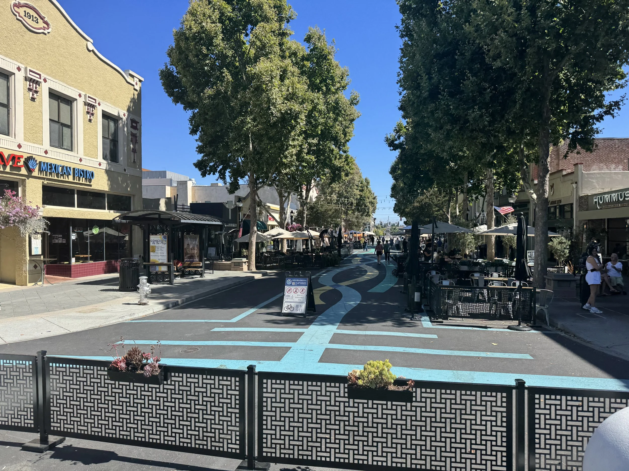 A sunny outdoor street scene with trees, outdoor seating for cafes, and pedestrians walking along the sidewalk and crosswalks.