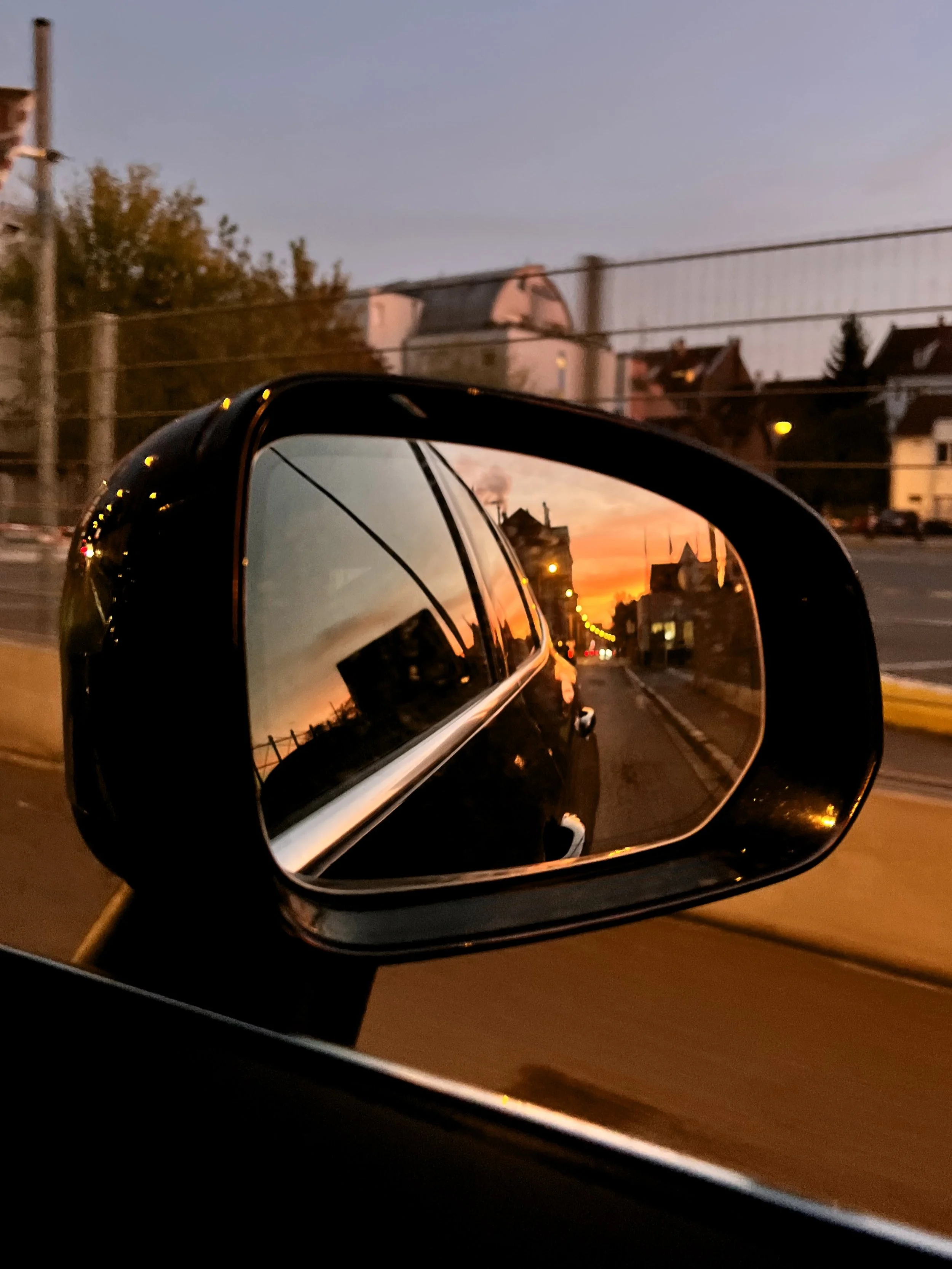 Reflet du coucher de soleil dans un rétroviseur de voiture sur une rue en ville avec des maisons et des arbres.
