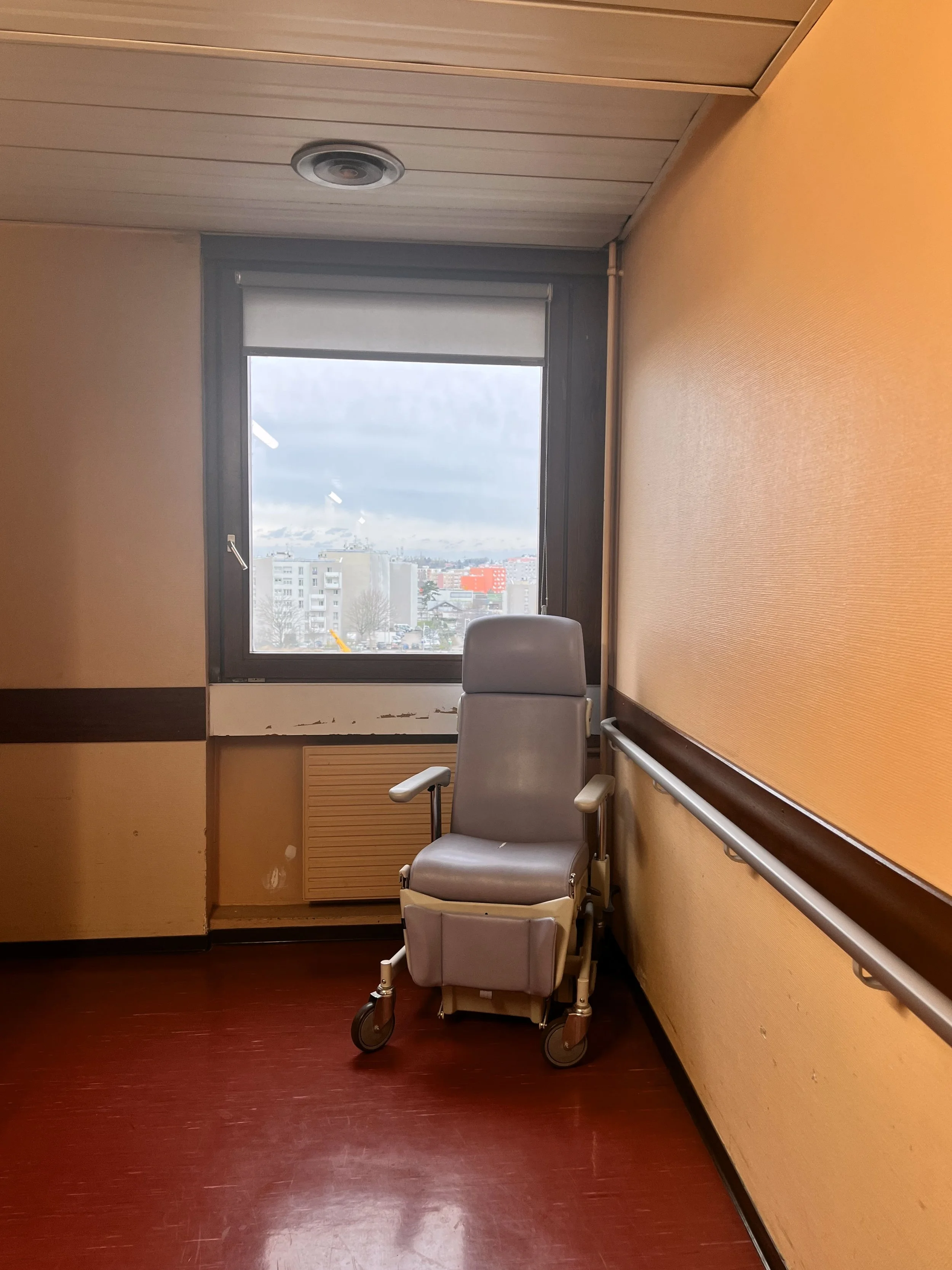 Salle d'hôpital avec un fauteuil roulant devant une grande fenêtre, murs orange et sol en bois foncé.
