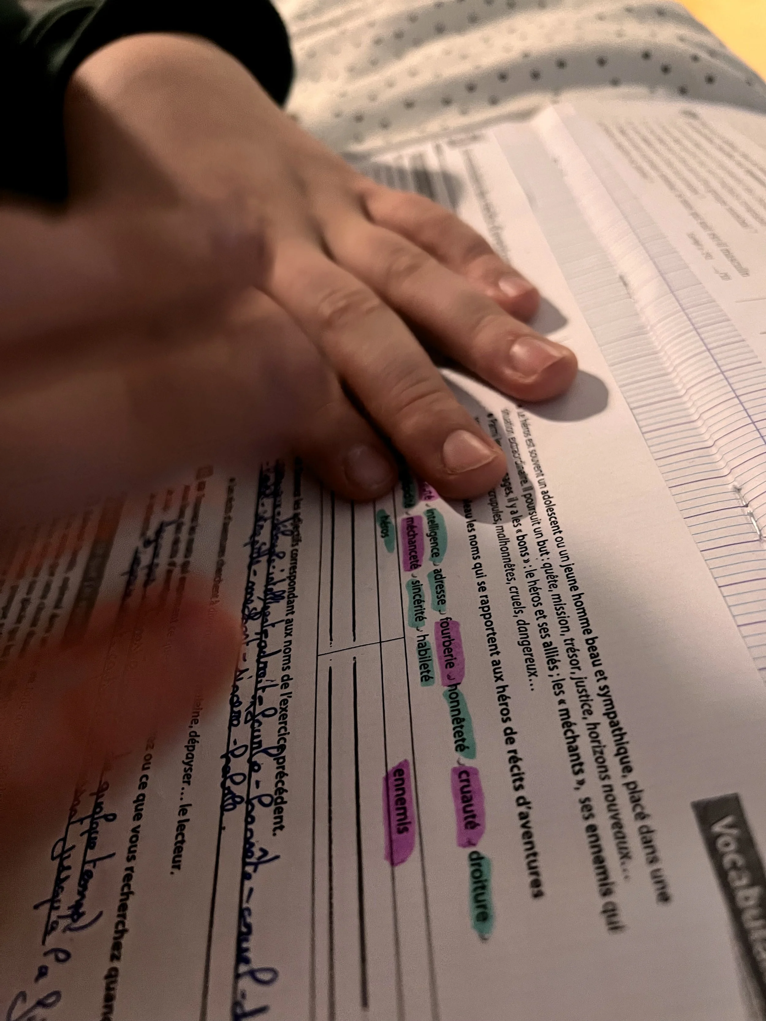 Une main posée sur une page de livre ou de cahier avec des mots soulignés et mis en évidence par des surligneurs de différentes couleurs.