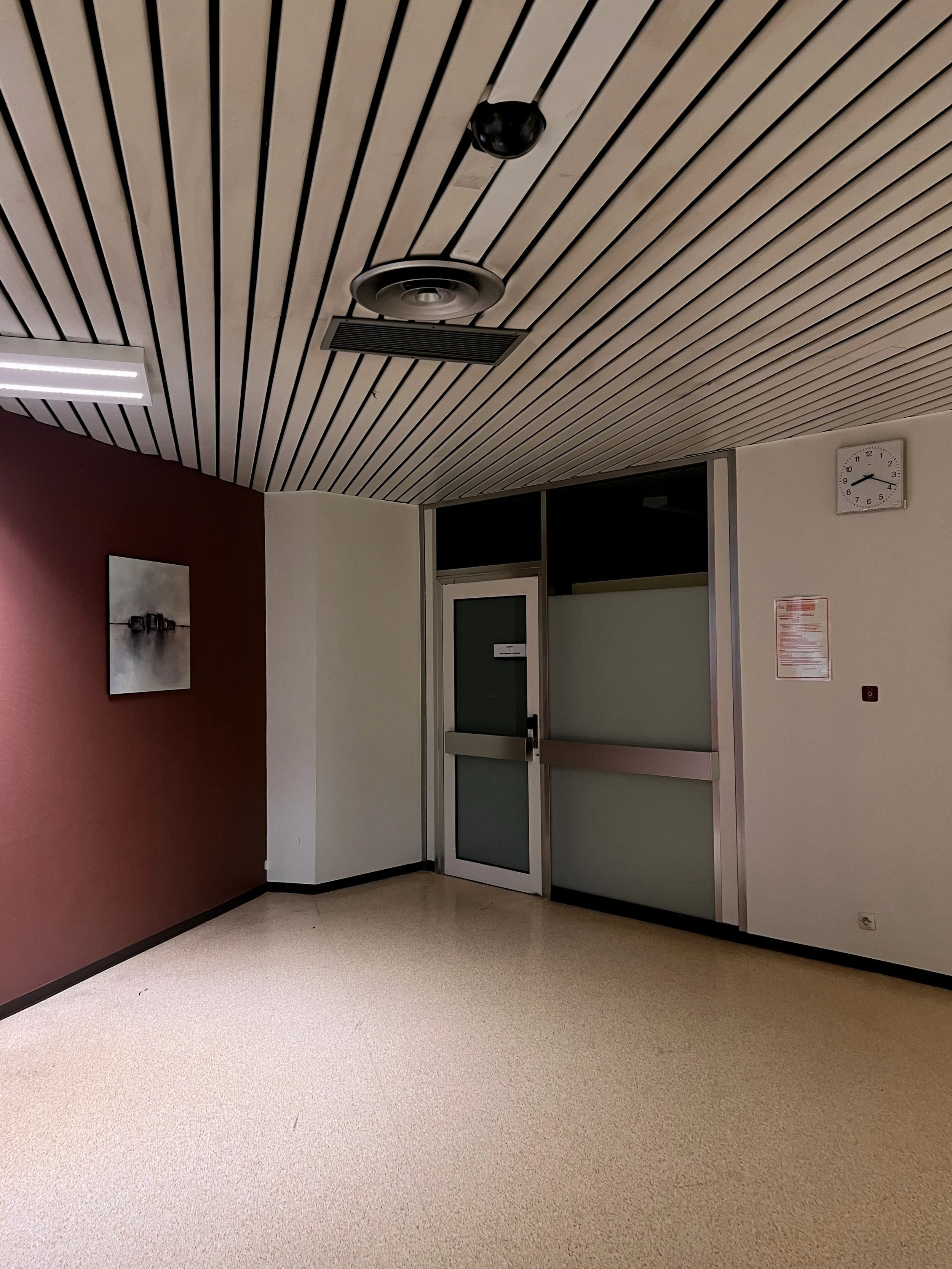 Intérieur d'un bâtiment avec mur blanc et mur rouge, porte en verre, horloge indiquant 3h14, tableau sur le mur rouge, plafond avec ventilation et caméra de surveillance, sol en moquette beige.