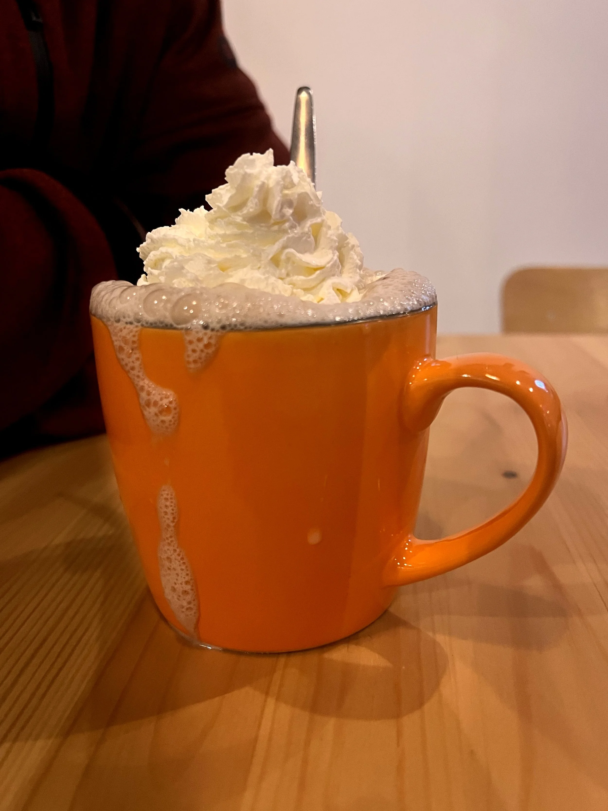 Une tasse orange remplie de chocolat chaud garni de crème fouettée avec une cuillère en argent à l'intérieur, posée sur une table en bois.