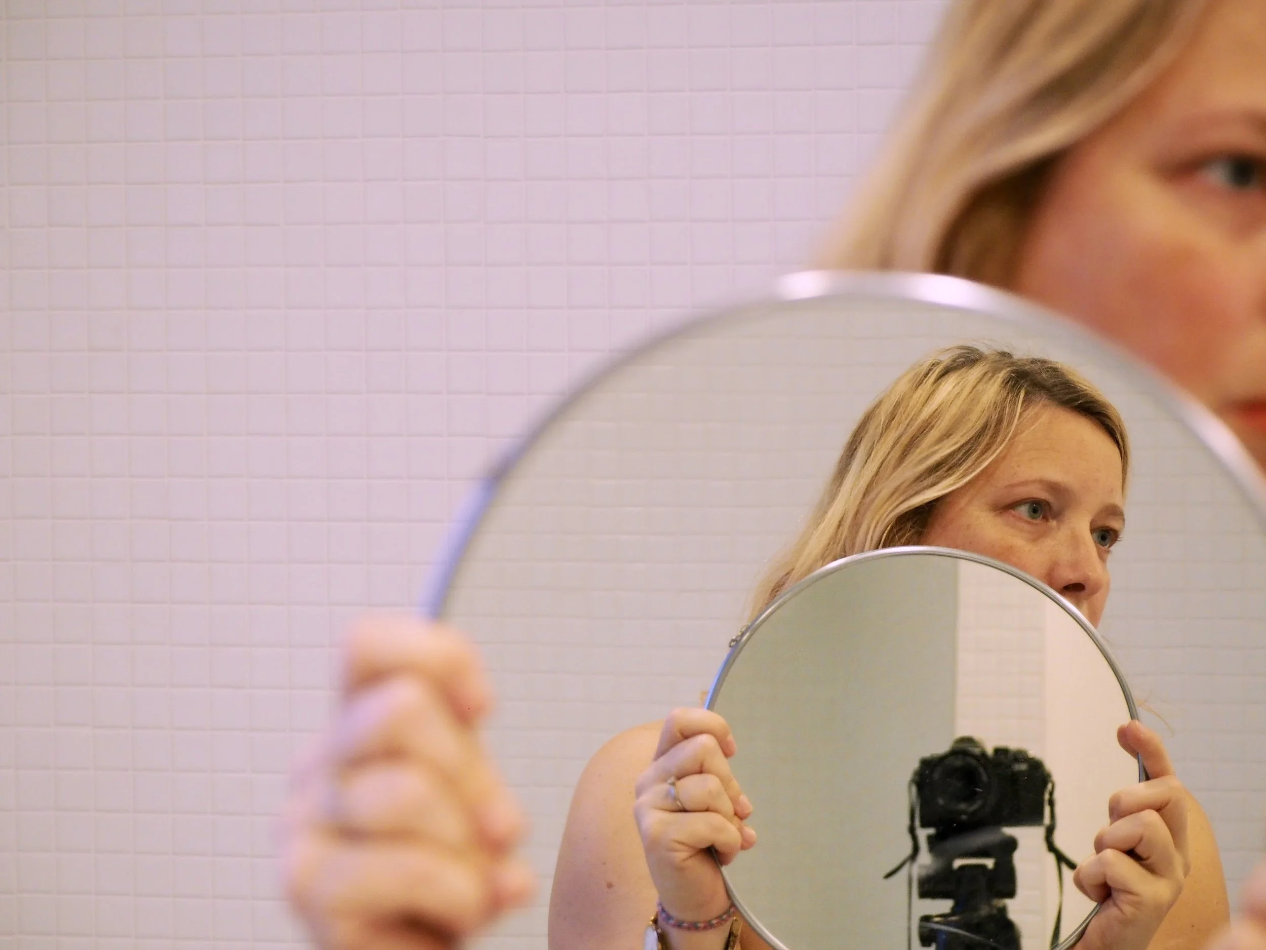 Une femme blonde tenant un miroir rond et se prenant en selfie avec un appareil photo noir, en arrière-plan un mur rose avec des carreaux.