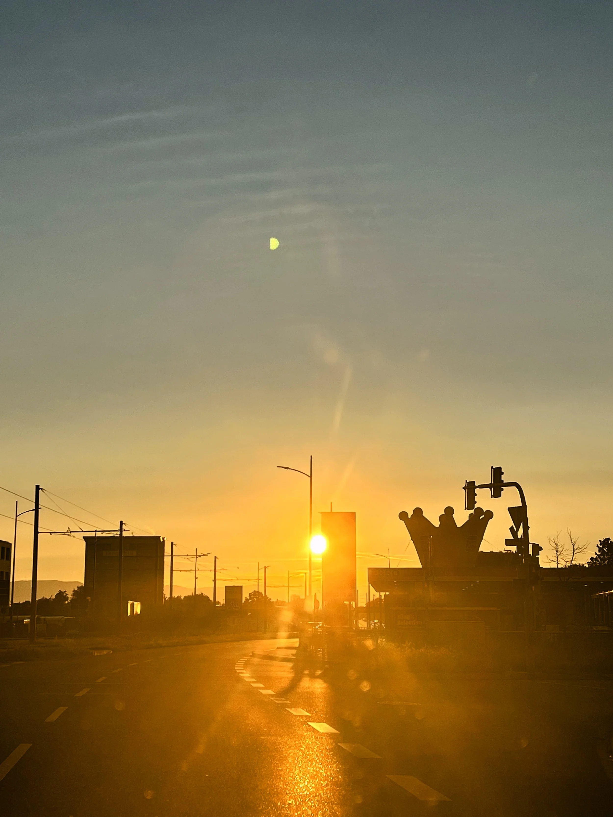 Un paysage urbain au coucher du soleil avec un ciel coloré allant du jaune à l'orange et au bleu, des silhouettes de bâtiments, de poteaux électriques et un feu de circulation. Le soleil est en position basse, proche de l'horizon, créant une lumière dorée sur la route.
