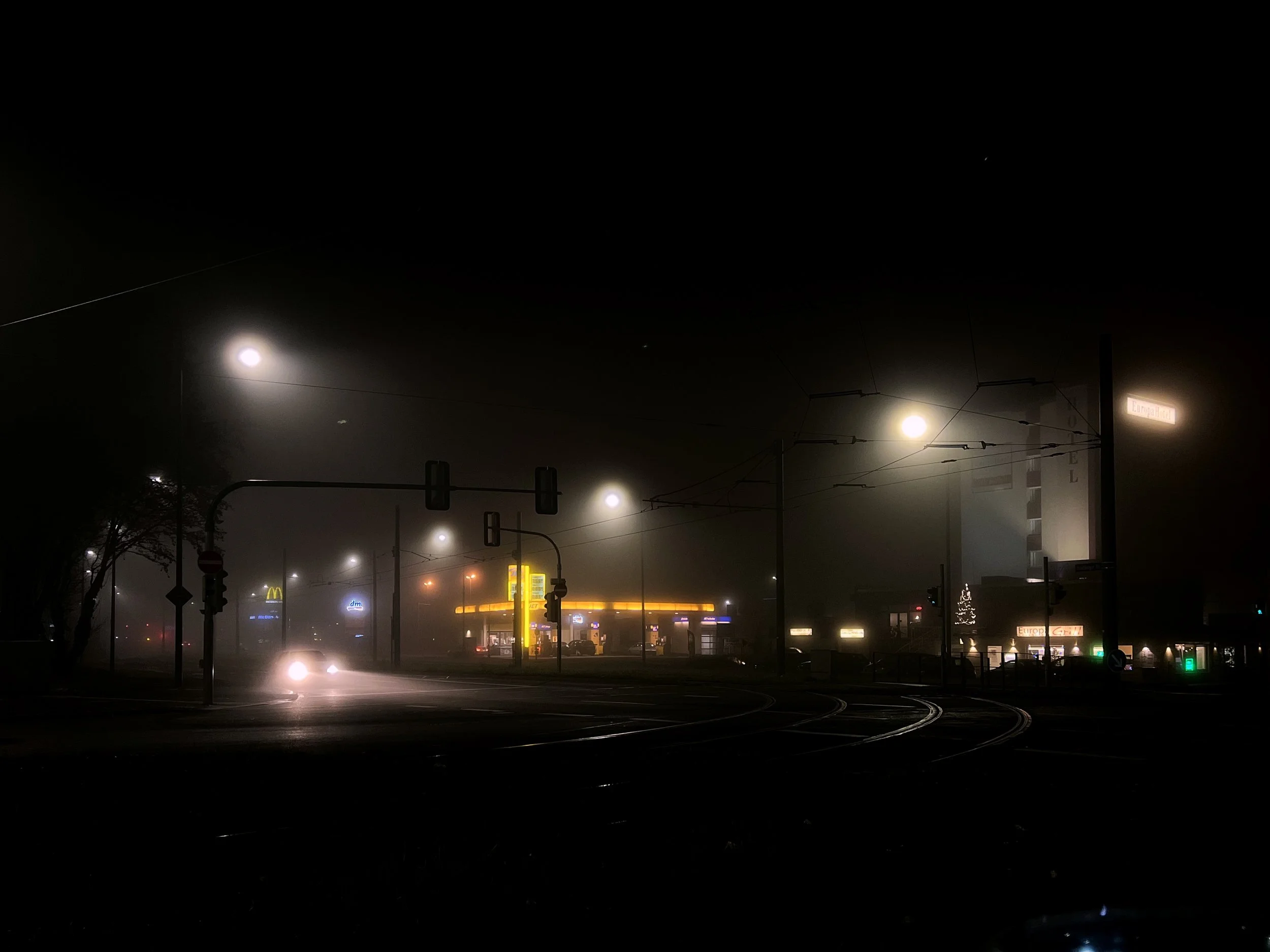 Une scène nocturne dans une ville brumeuse avec des lampadaires illuminant la rue, des bâtiments, et des panneaux lumineux, notamment une enseigne "Europalace" et un logo de McDonald's en arrière-plan.