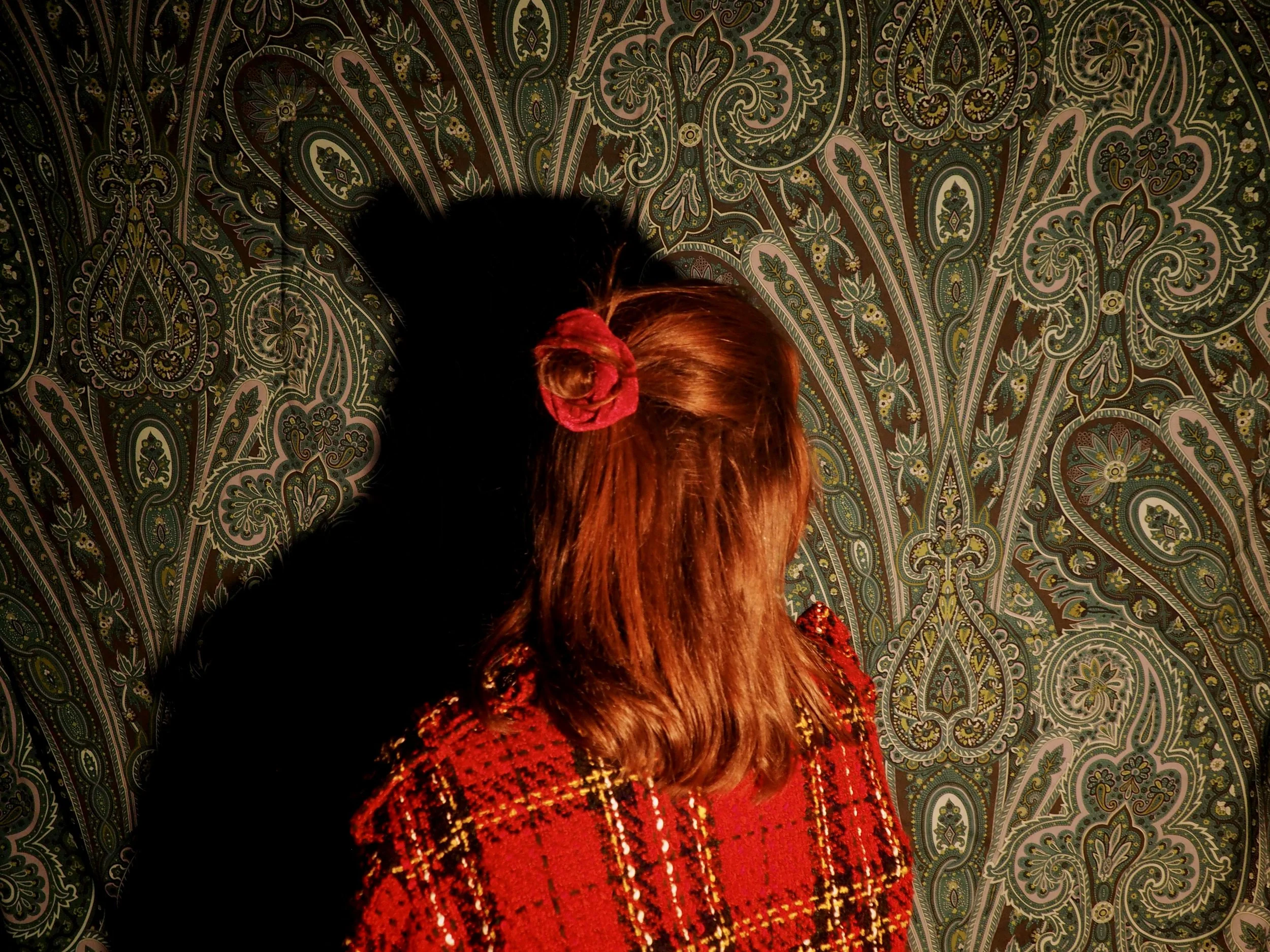 Une fille avec des cheveux roux, portant un ruban rouge dans ses cheveux, dans une pièce avec un papier peint à motif floral complexe.