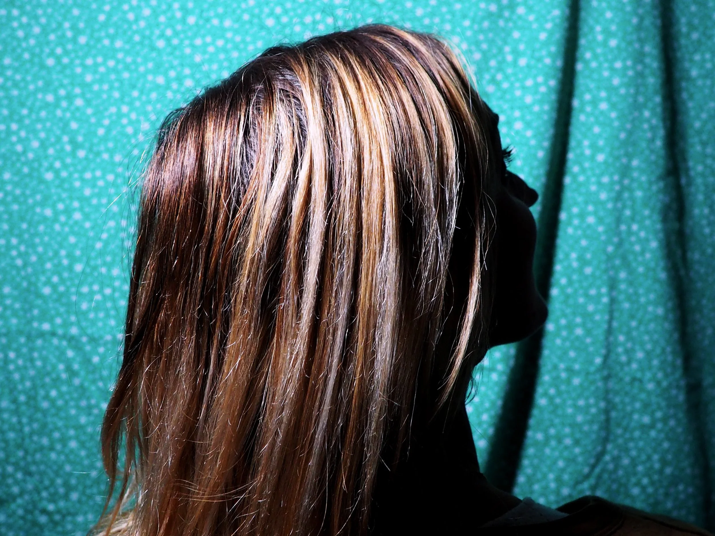 Cheveux longs, lisses, avec reflets blondes, brun et cuivres, sur une personne dont le visage n'est pas visible, devant un fond vert à pois blancs.