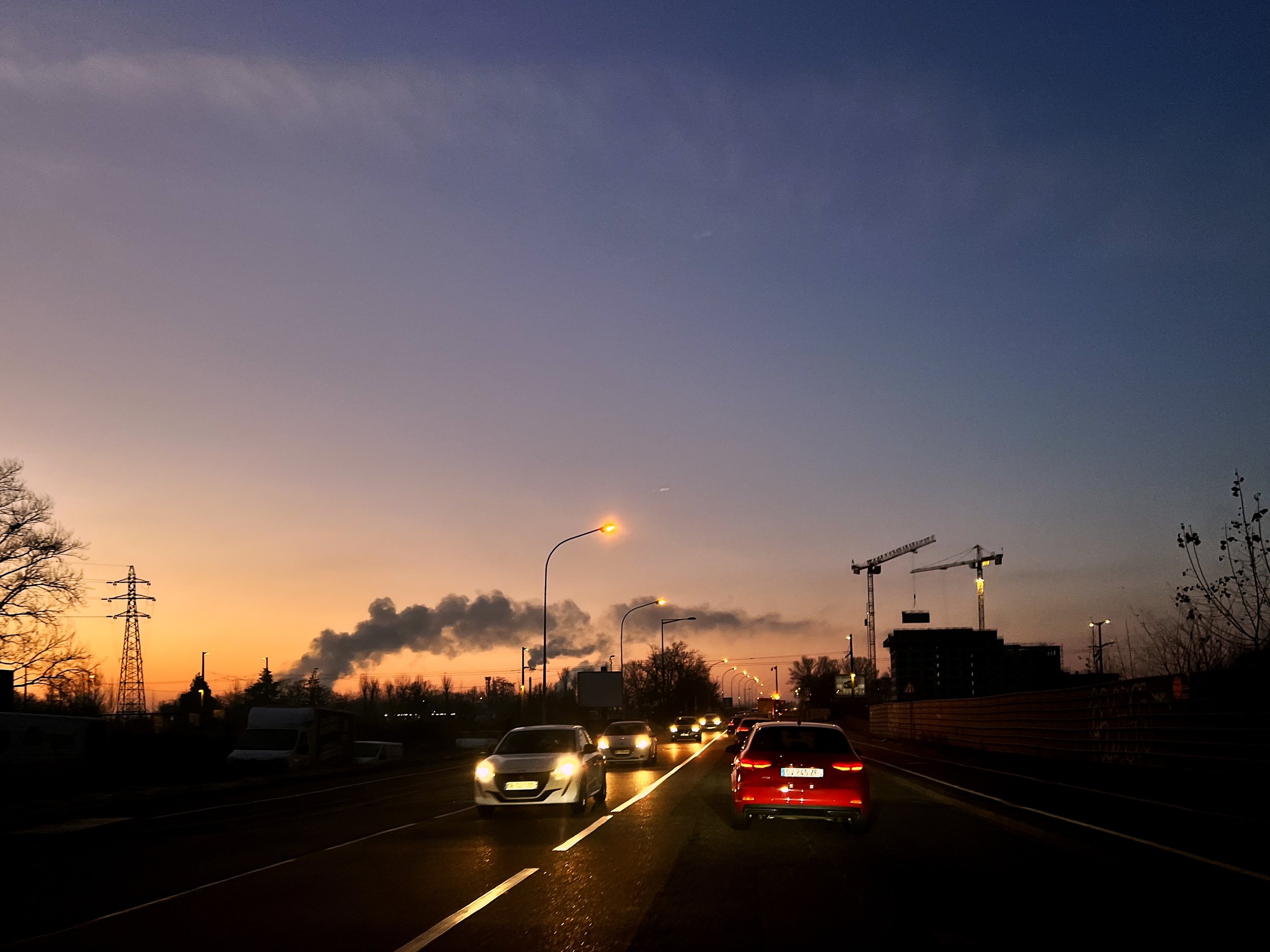 Coucher de soleil derrière une ville avec des voitures sur une route, des lampadaires allumés et des grues de construction.