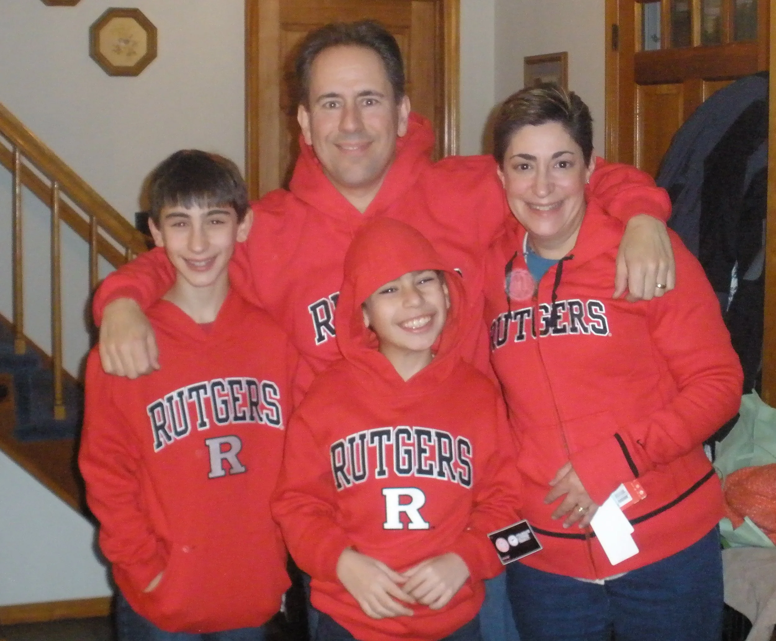 Group of four people wearing red 'Rutgers' sweatshirts, with three children and an adult, standing inside a home. The background features a staircase and wooden cabinets.