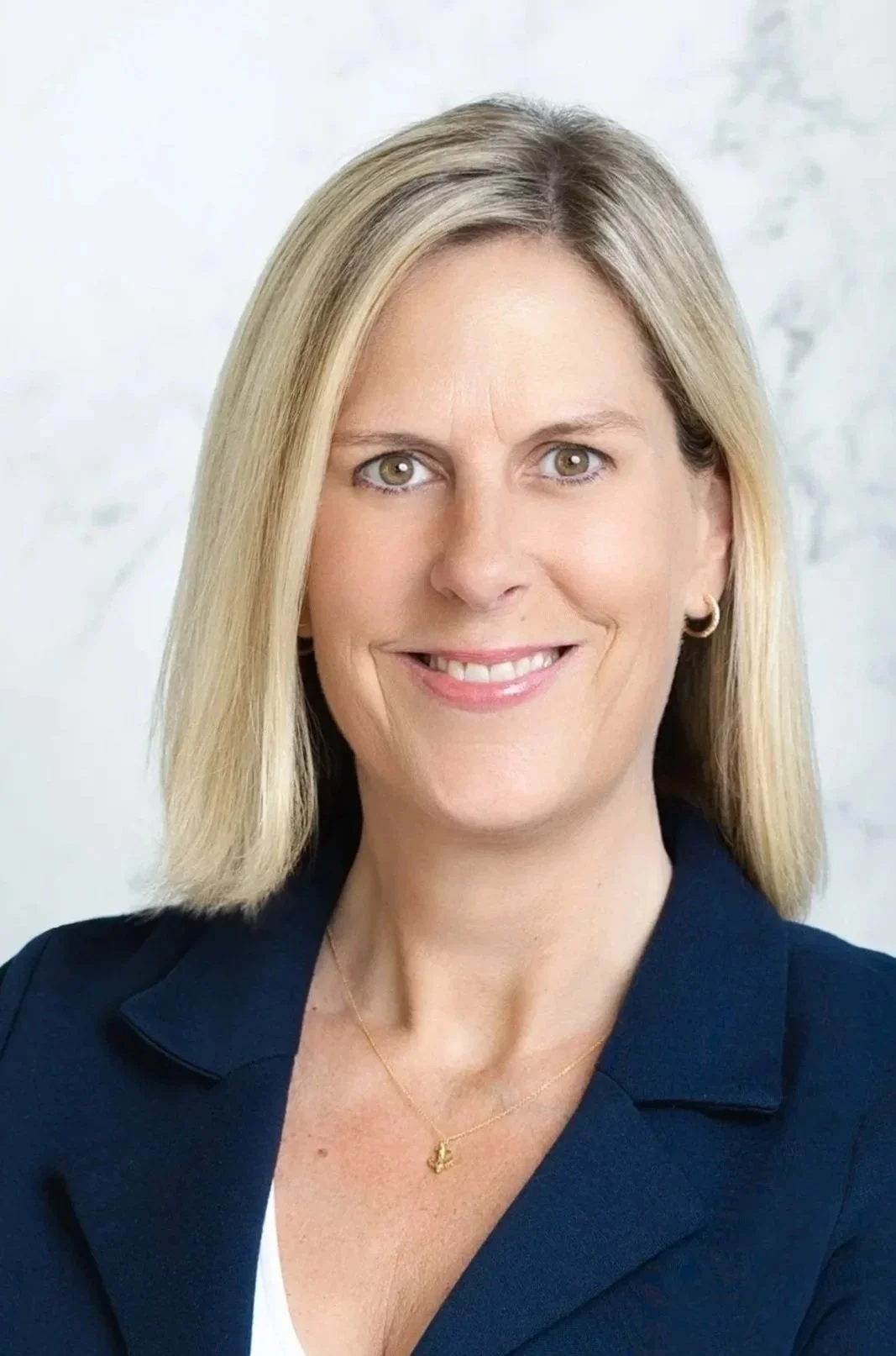 A woman with blonde hair, wearing a navy blazer, gold hoop earrings, and a gold necklace, smiling at the camera with a light-colored background.