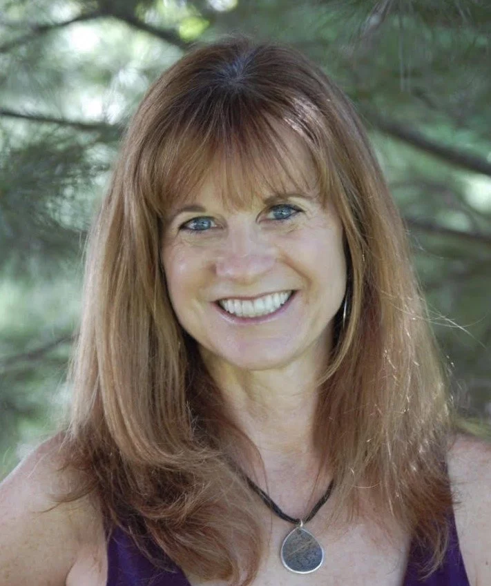 A woman with shoulder-length red hair and blue eyes smiling outdoors, wearing a purple top and a black necklace with a silver pendant.
