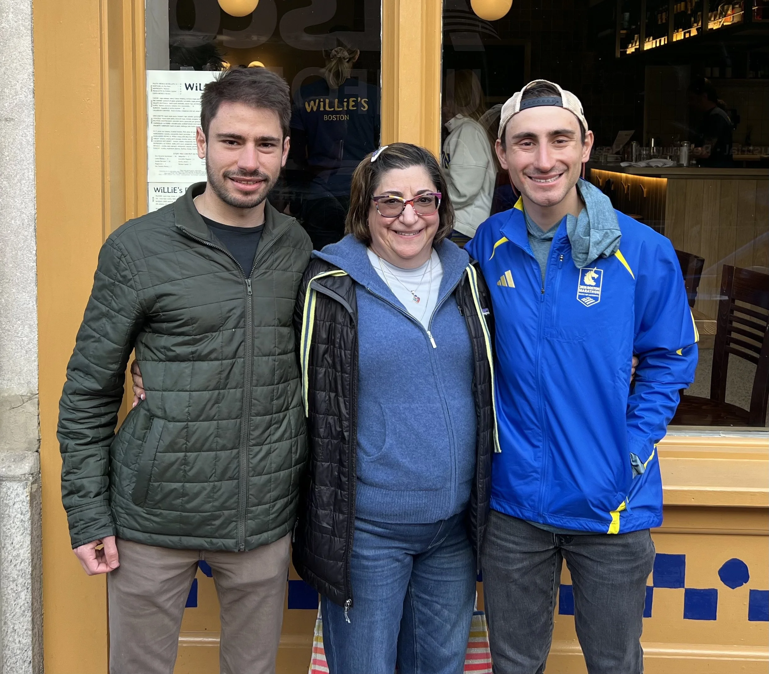 Three people standing together outside a restaurant with yellow window trim, smiling at the camera.