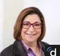 A woman with shoulder-length hair, wearing glasses and a black blazer over a purple shirt, smiling at the camera.