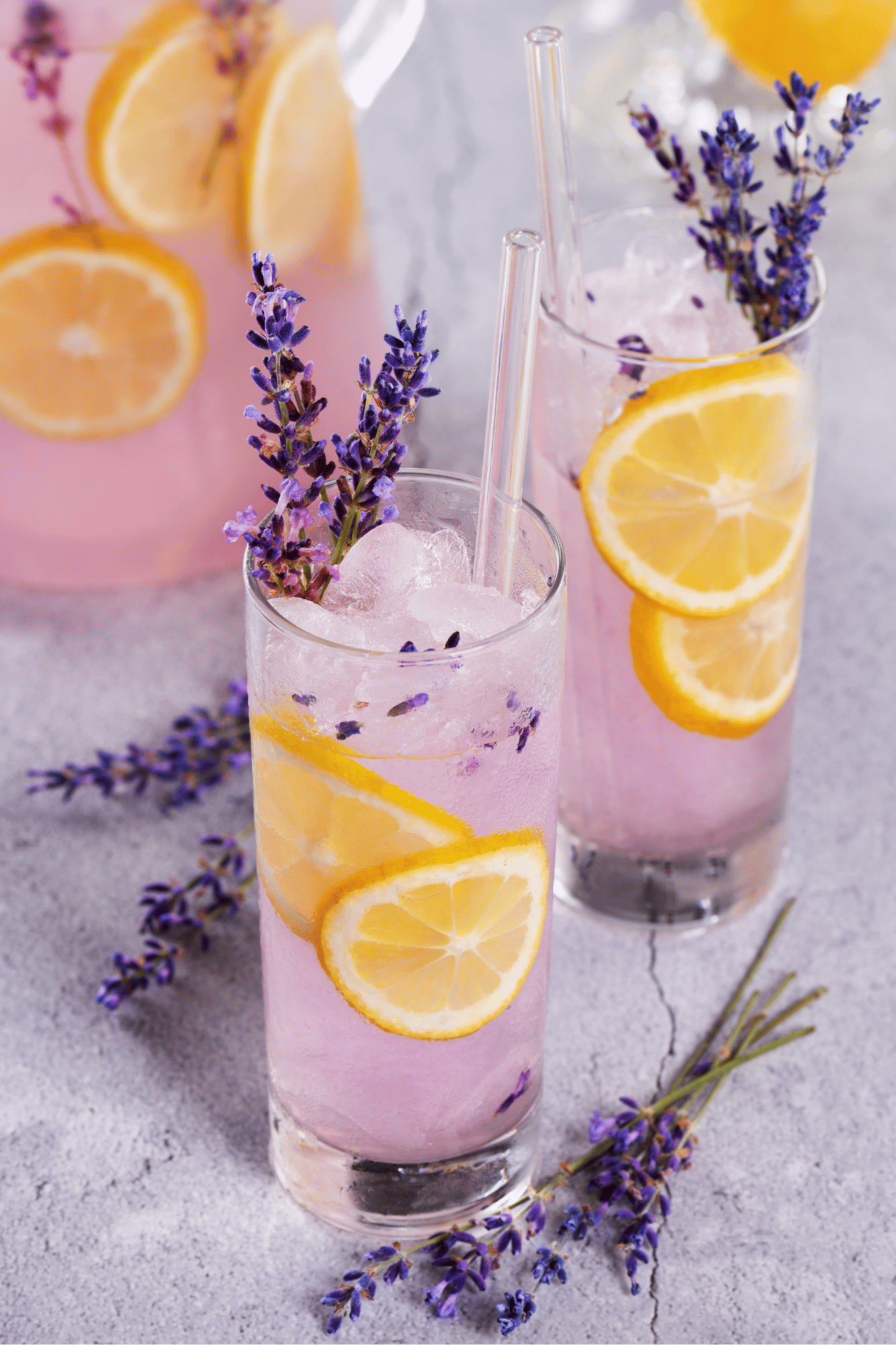 Two tall glasses of pink lemonade with slices of lemon and sprigs of lavender, set on a textured surface with scattered lavender sprigs in the background.