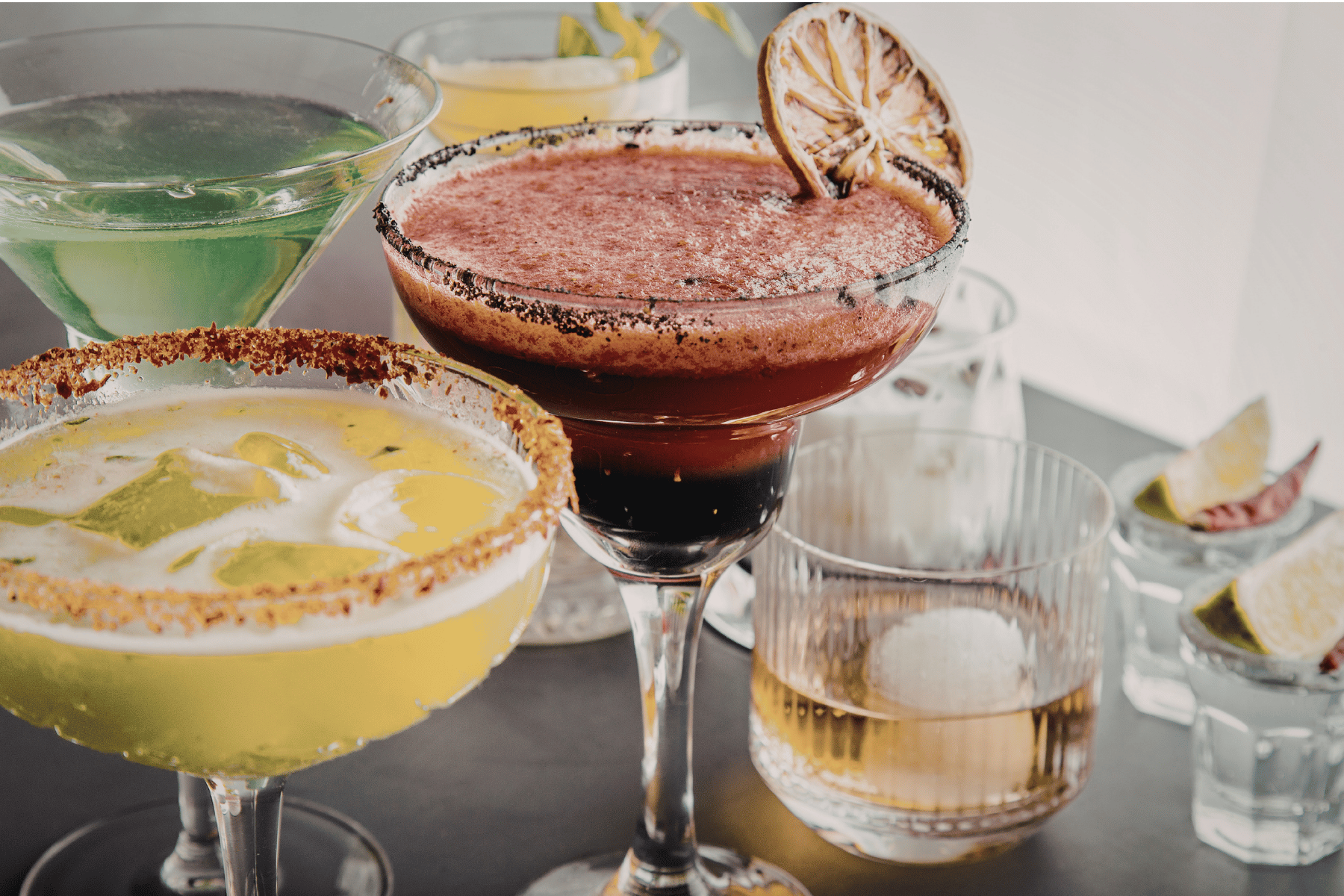 Various cocktails including a green drink, a yellow drink with a lemon slice, a red frozen drink with a dried citrus garnish, and a clear drink with ice and a slice of lime, all displayed on a bar or table setting.