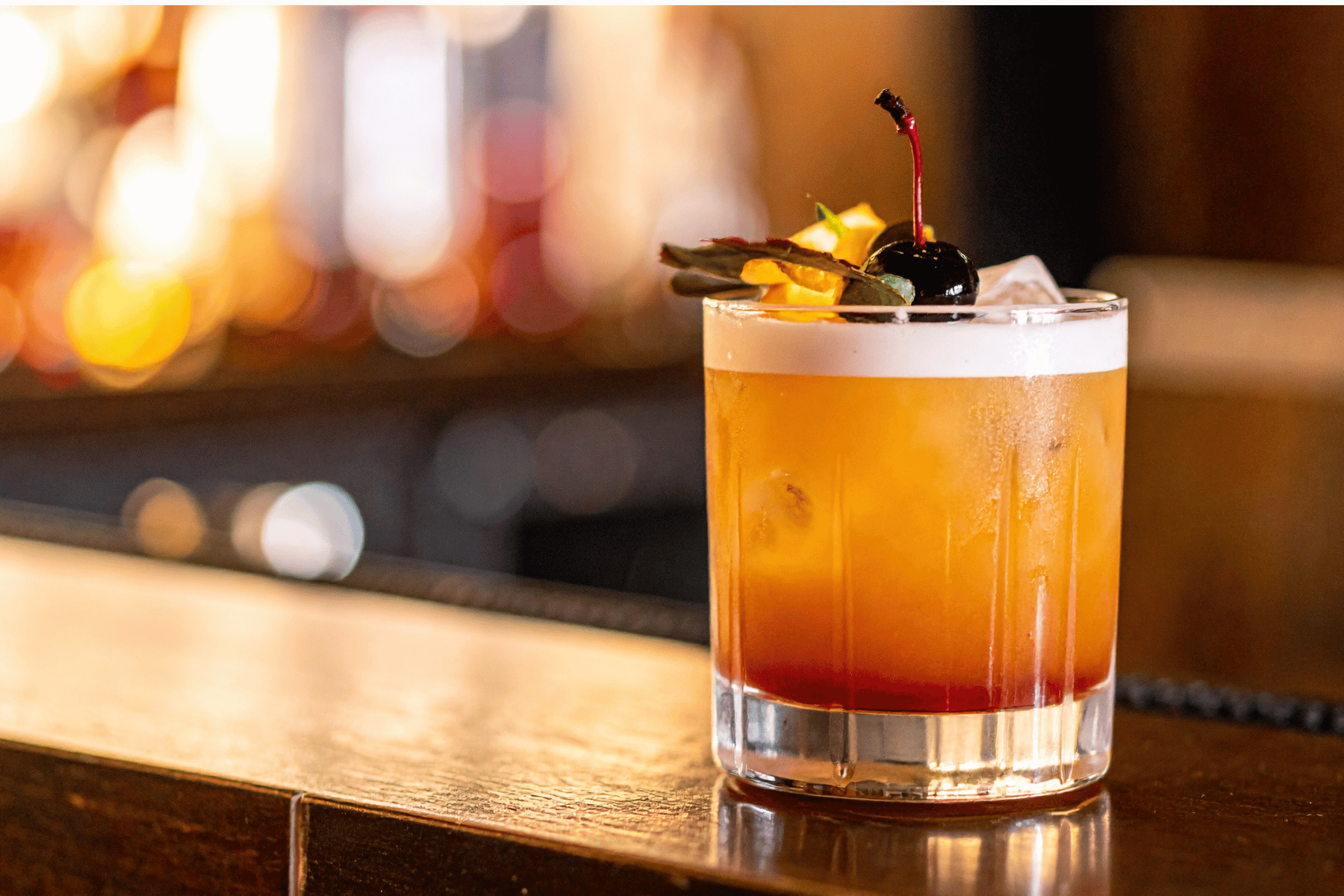 A cocktail in a short glass garnished with a dark cherry, lime peel, and a cherry stem, set on a wooden bar with colorful blurred lights in the background.