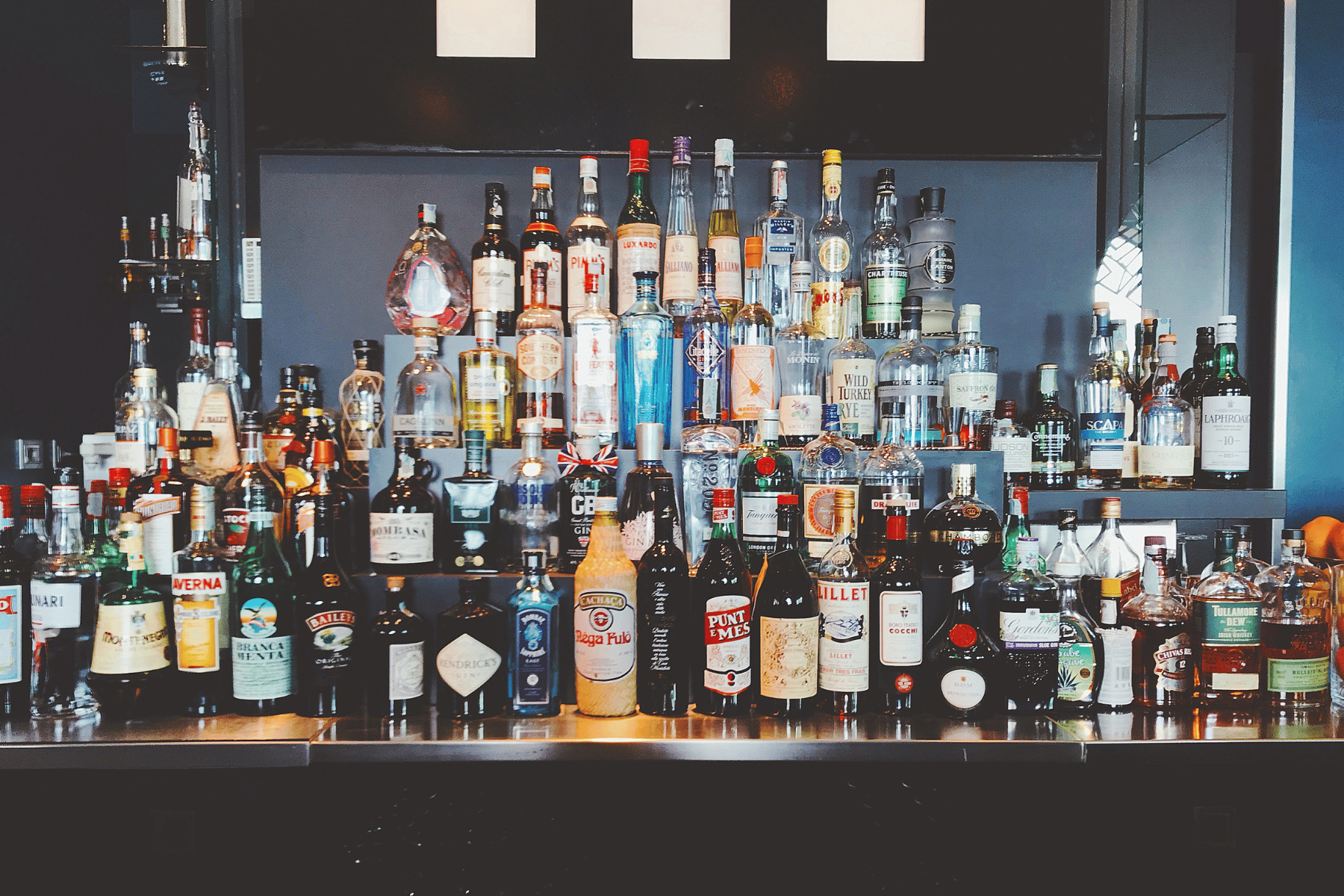 A black bar shelf filled with various bottles of liquor, including whiskey, gin, and vodka, arranged in front of a black wall with three white rectangular panels.