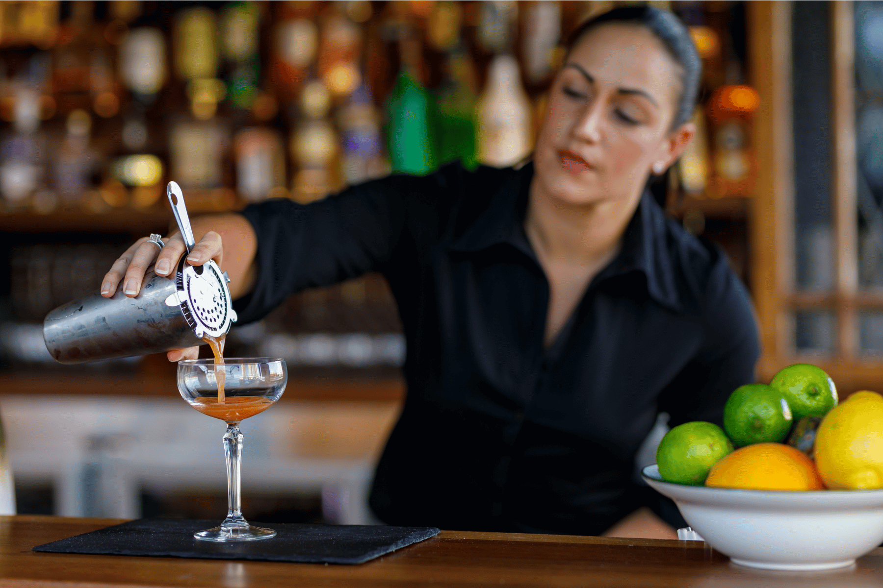 A woman pouring a cocktail from a shaker into a glass at a bar. There are limes, lemons, and a bowl of citrus fruits on the counter.