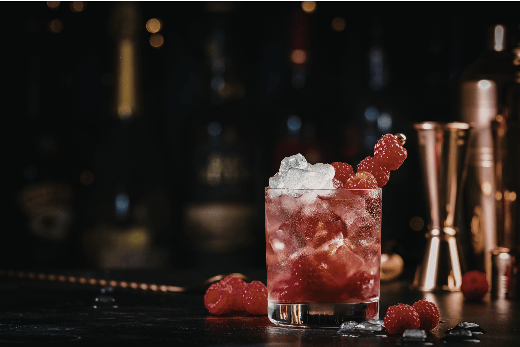 A glass of raspberry cocktail with ice cubes and fresh raspberries on top, on a dark bar counter with scattered raspberries and ice pieces, with blurred bar tools in the background.