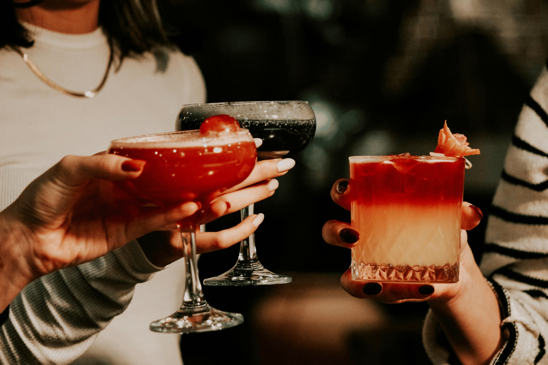 Three women are holding different cocktails in glasses at a social gathering, raising them together in a toast.
