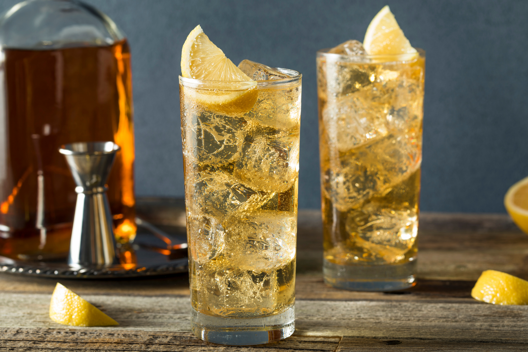 Two tall glasses filled with a sparkling yellow beverage and ice, garnished with lemon wedges, on a wooden surface, with a liquor decanter and metal bartender tools in the background.