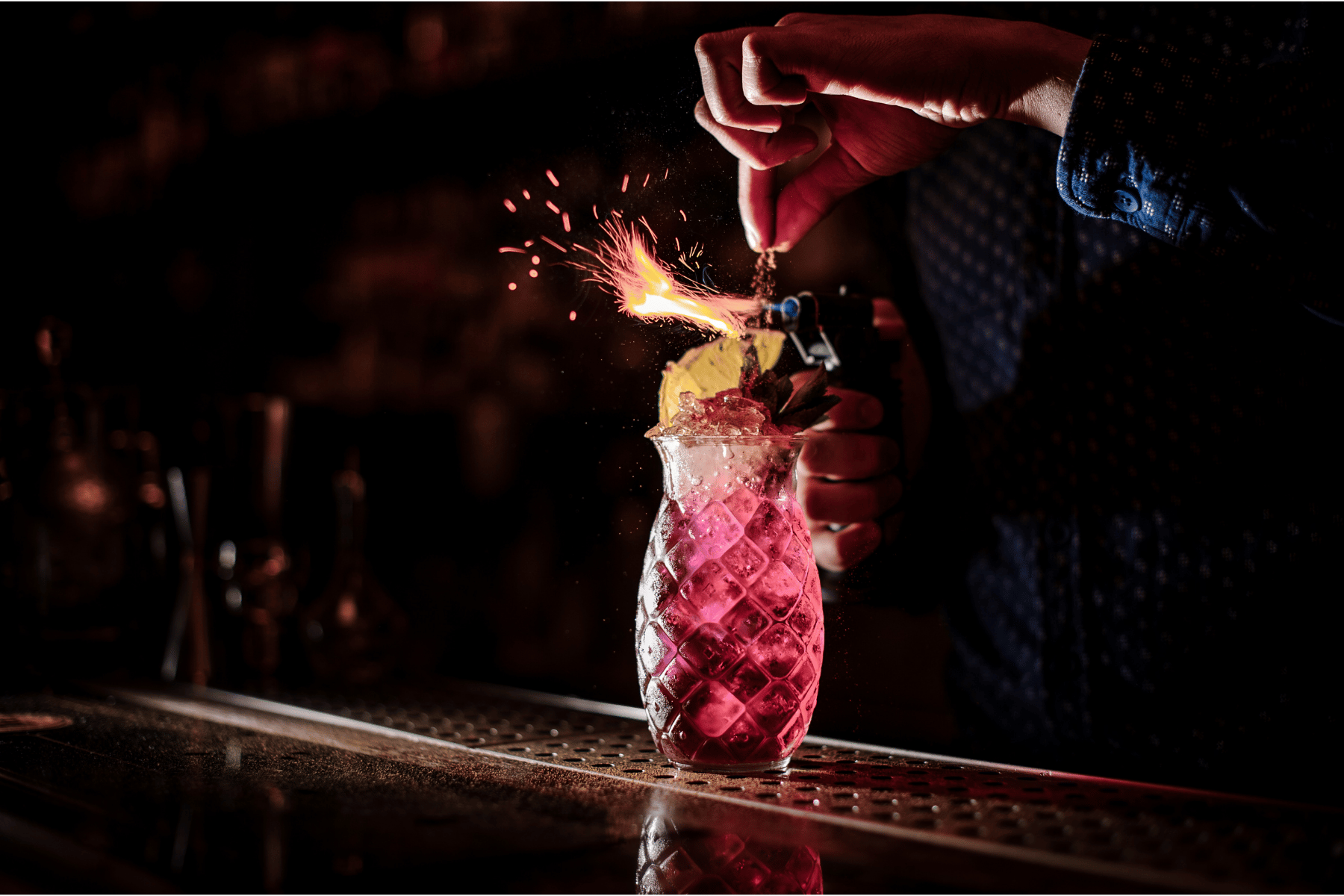 A person is igniting a firework in a decorative glass filled with a pink beverage, with sparks flying out of the firework.