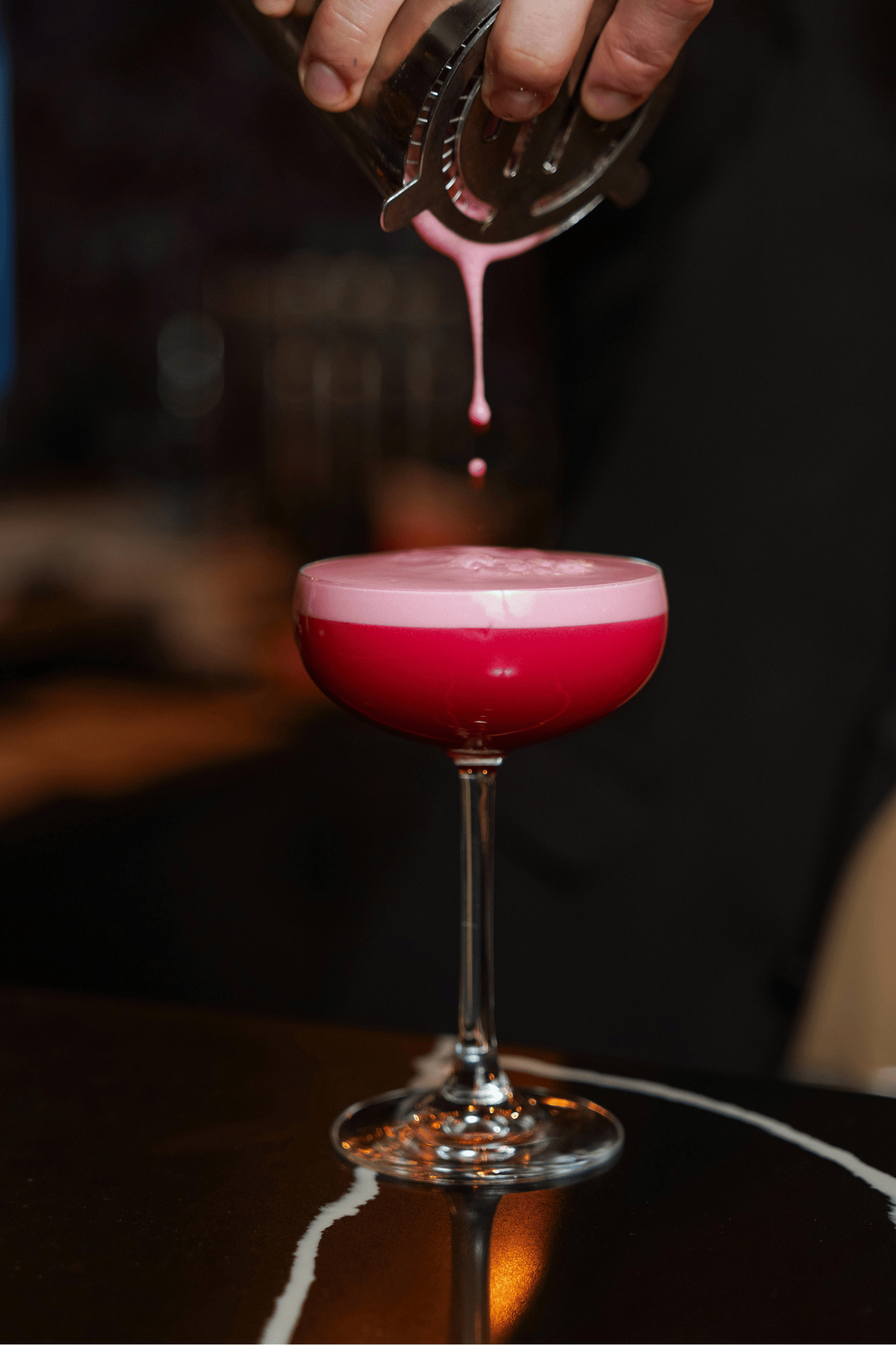 A pink cocktail in a coupe glass being poured by a bartender, with the drink overflowing slightly. The scene is dark and elegant, with the drink's reflection visible on the table.