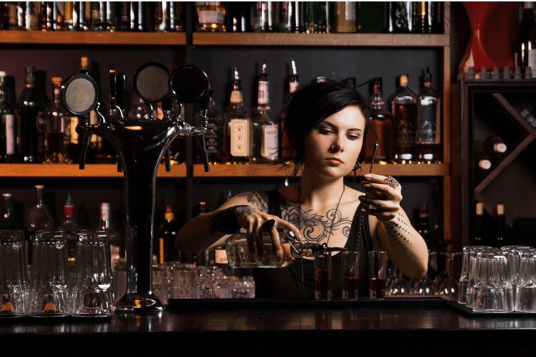 A woman with tattoos and dark hair behind a bar, pouring a red cocktail into a glass, with various bottles and glasses on shelves and counter.